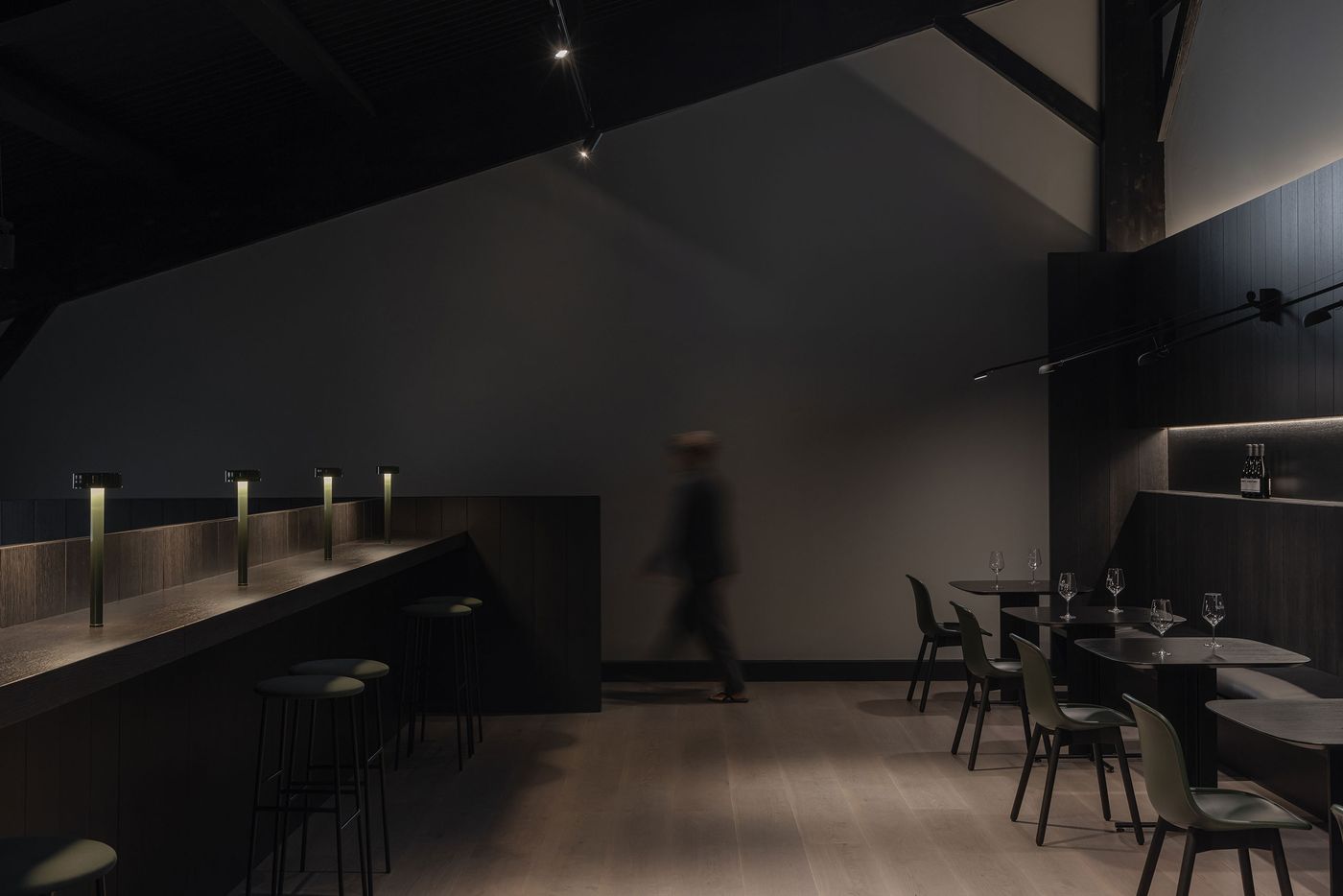 A dimly lit modern restaurant interior with stools at a bar, tables set with glasses, and a blurred figure walking by. A dimly lit modern restaurant interior with stools at a bar, tables set with glasses, and a blurred figure walking by.