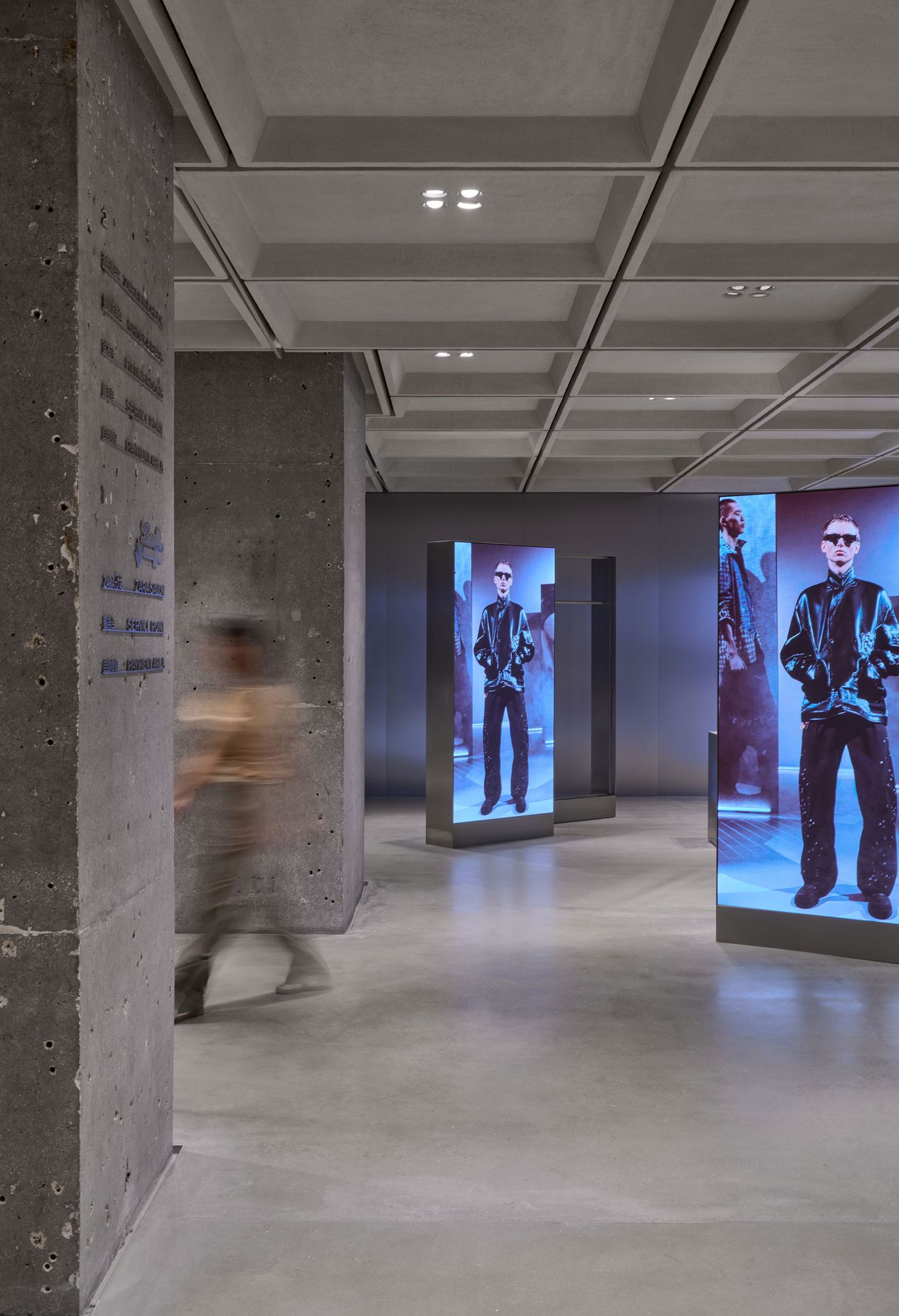 A modern exhibition space featuring digital displays of fashion pieces, with gray concrete walls and a sleek design. A visitor walks through.