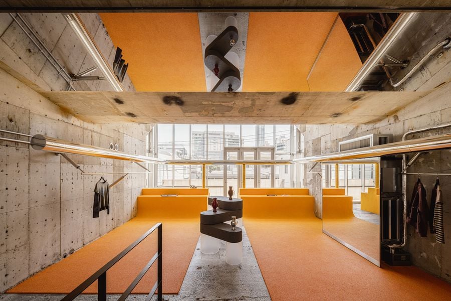 A wide view of the top level shows I IN’s restrained retail choreography: vivid yellow flooring and low integrated displays set against rough concrete, with chrome rails tracing both sides of the room. A central sculptural fixture and mirrored wall organise the space while preserving a sense of openness towards the glazed façade.