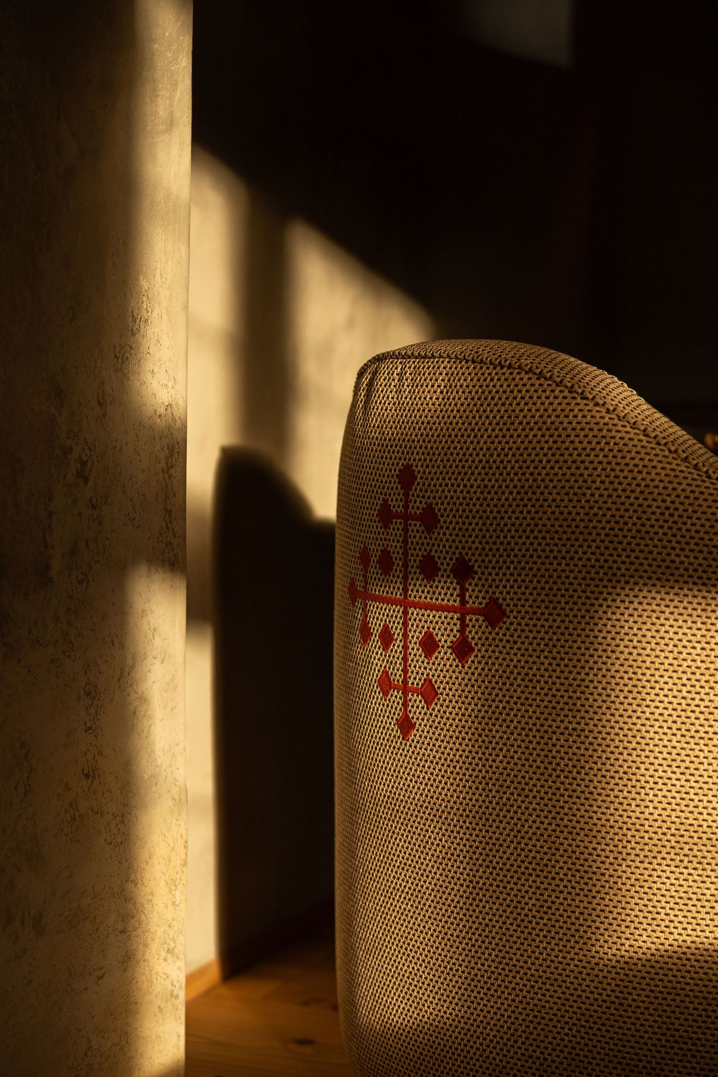 A close study of texture and light at Castel Badia: a cross motif embroidered in rust thread on the back of a woven upholstered chair. A slanting shaft of sunlight and the shadow of a pillar fall across a mottled plaster wall and pine floorboards beside it. A close study of texture and light at Castel Badia: a cross motif embroidered in rust thread on the back of a woven upholstered chair. A slanting shaft of sunlight and the shadow of a pillar fall across a mottled plaster wall and pine floorboards beside it.