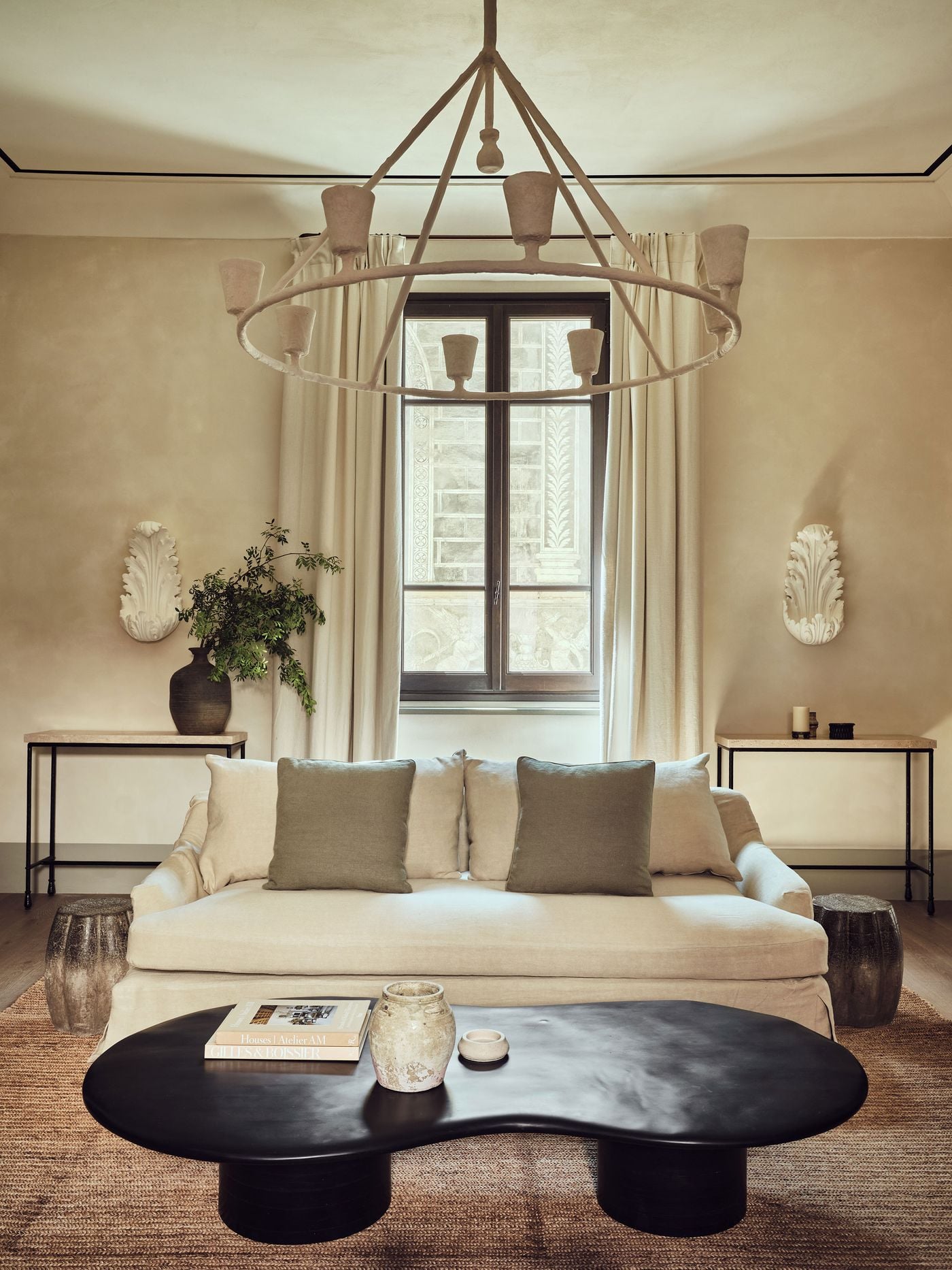 A ring chandelier of plaster cups hovers above a slipcovered linen sofa in a living room at La Réserve Firenze, facing a biomorphic black coffee table on a jute rug. White acanthus wall ornaments, stone-topped iron consoles and a window onto a sgraffito facade complete the restrained, sculptural scene. A ring chandelier of plaster cups hovers above a slipcovered linen sofa in a living room at La Réserve Firenze, facing a biomorphic black coffee table on a jute rug. White acanthus wall ornaments, stone-topped iron consoles and a window onto a sgraffito facade complete the restrained, sculptural scene.
