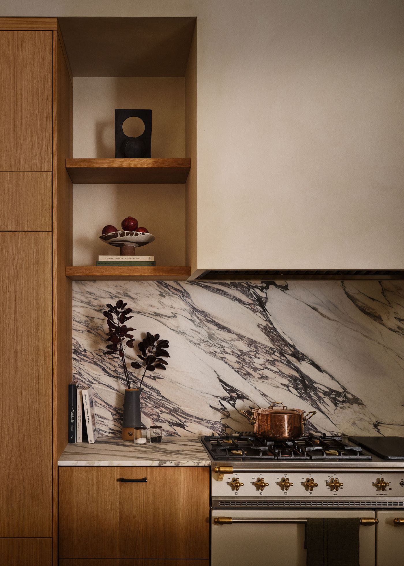 A modern kitchen featuring marble countertops, a gas stove, copper pot, shelf decor, and elegant dark foliage in a vase.