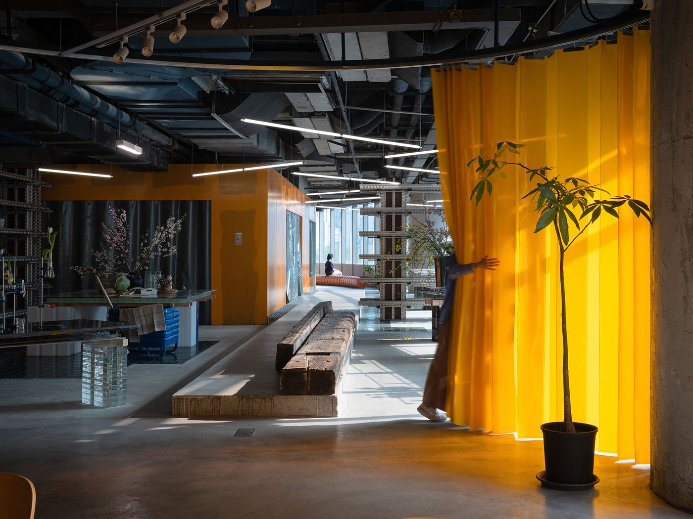 A person gently interacts with a voluminous yellow curtain in 889 GLO Art Space, highlighting its role as a flexible, colorful partition within the industrial setting. The raw concrete floors and ceiling are balanced by a long wooden bench and elegant floral arrangements, creating a vibrant yet contemplative atmosphere. A person gently interacts with a voluminous yellow curtain in 889 GLO Art Space, highlighting its role as a flexible, colorful partition within the industrial setting. The raw concrete floors and ceiling are balanced by a long wooden bench and elegant floral arrangements, creating a vibrant yet contemplative atmosphere.