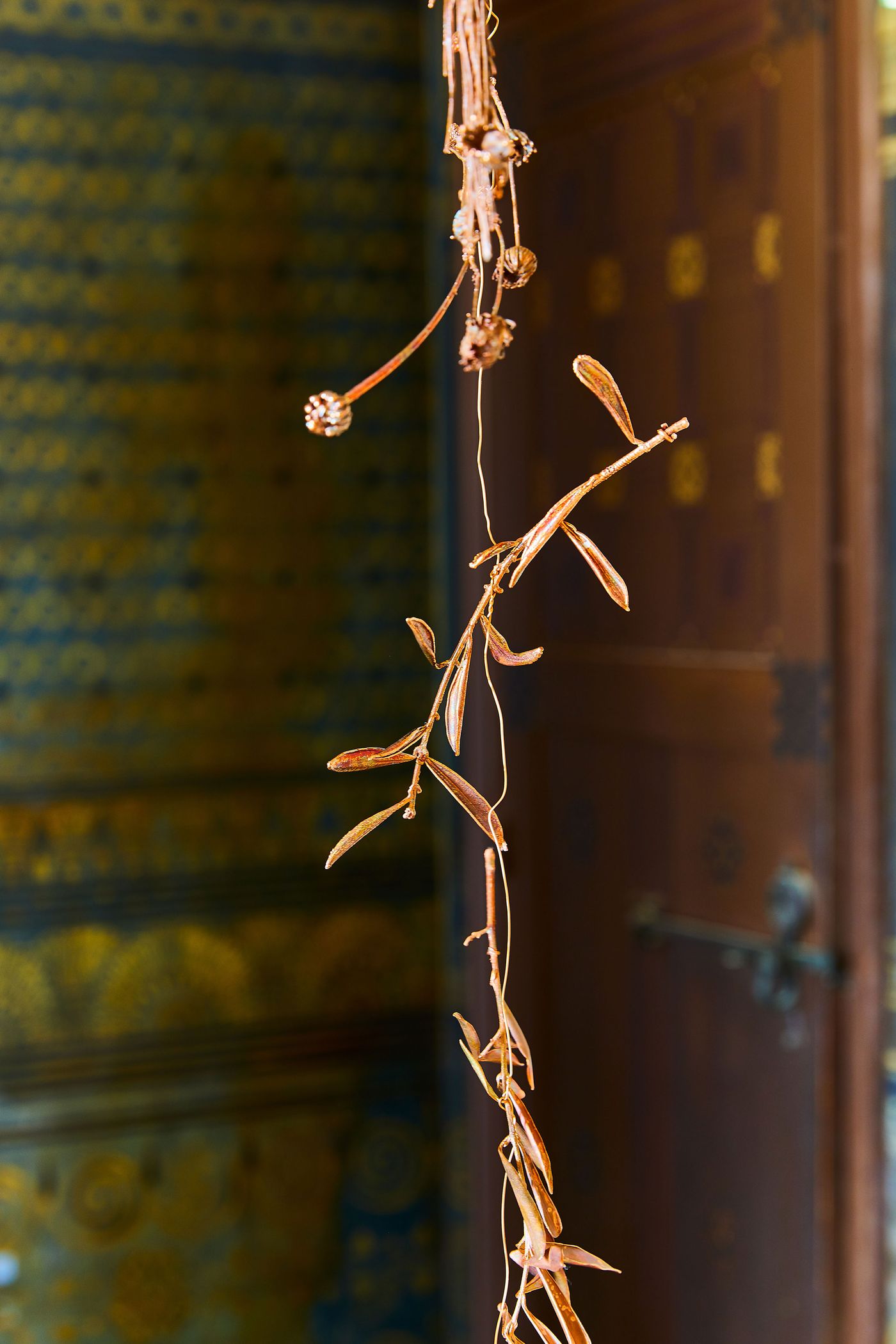 A close-up of a hanging art piece by Gabriel Leger, featuring a single, delicate branch with metallic leaves. The piece dangles in front of a richly textured wall with a blue and gold pattern and a dark, ornate wooden door with brass details. The juxtaposition of the fine, biophilic sculpture and the solid architecture creates a sense of fragility and strength.