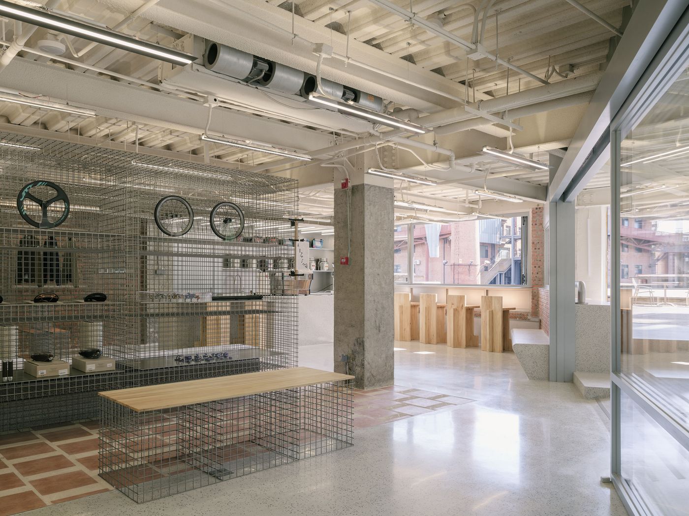 An expansive view of AITASHOP illustrates its multi-functional design, blending retail with communal spaces. Wire mesh display units for bike accessories integrate with a minimalist, stepped seating area, leading to a bright cafe zone with light wooden stools. The interplay of concrete, exposed brick, and terracotta tiles creates a layered, industrial-chic aesthetic, enhanced by generous natural light. An expansive view of AITASHOP illustrates its multi-functional design, blending retail with communal spaces. Wire mesh display units for bike accessories integrate with a minimalist, stepped seating area, leading to a bright cafe zone with light wooden stools. The interplay of concrete, exposed brick, and terracotta tiles creates a layered, industrial-chic aesthetic, enhanced by generous natural light.