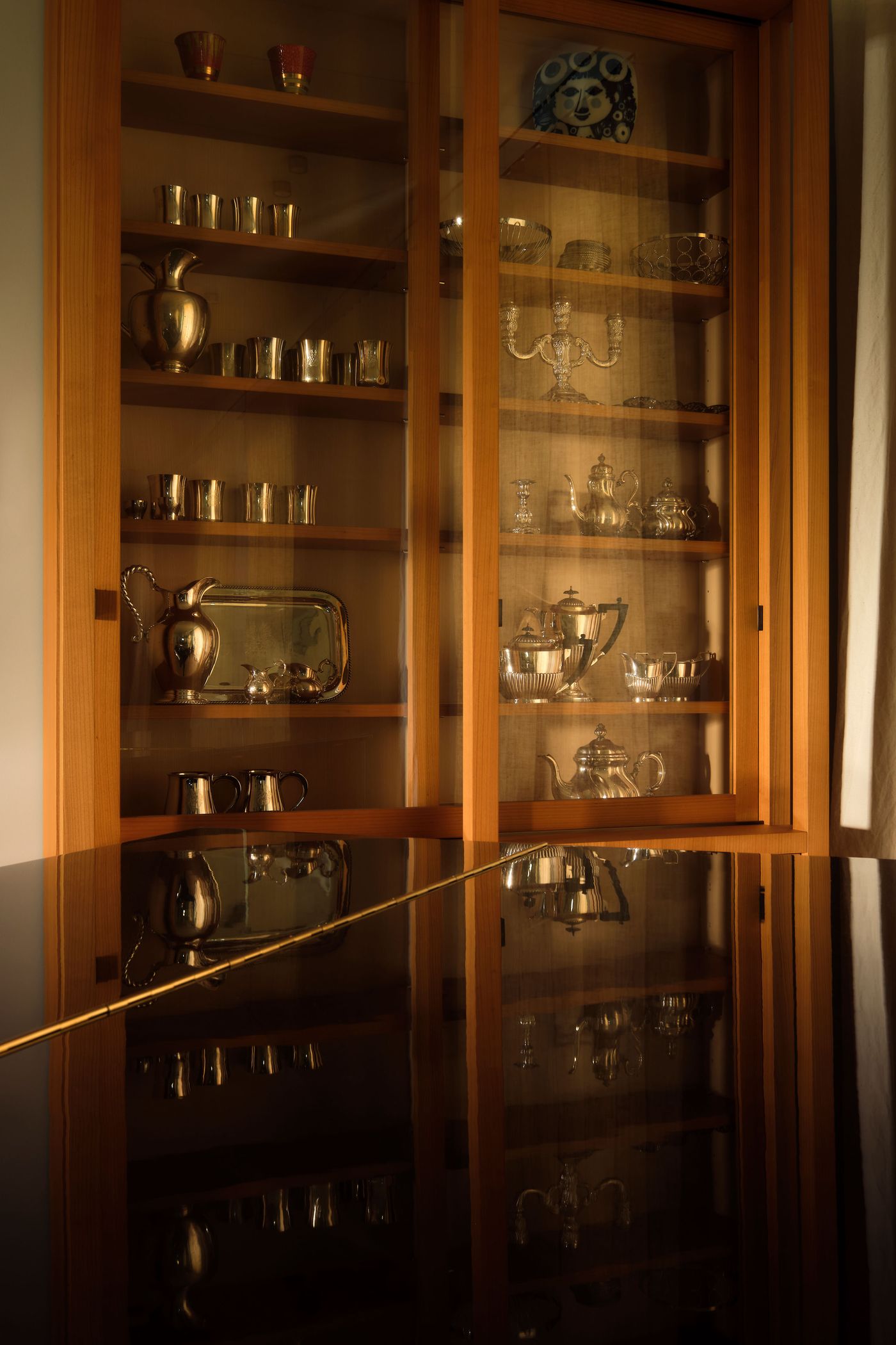 A wooden display cabinet showcases an array of silverware and glass pieces reflecting on a polished black table.