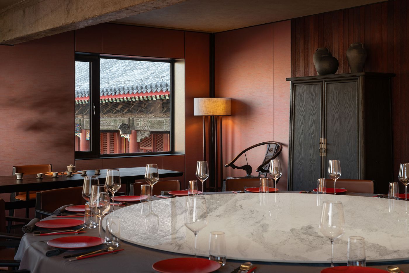 Elegant dining room featuring a round marble table set with red plates and glasses, overlooking traditional architecture through large windows. Elegant dining room featuring a round marble table set with red plates and glasses, overlooking traditional architecture through large windows.