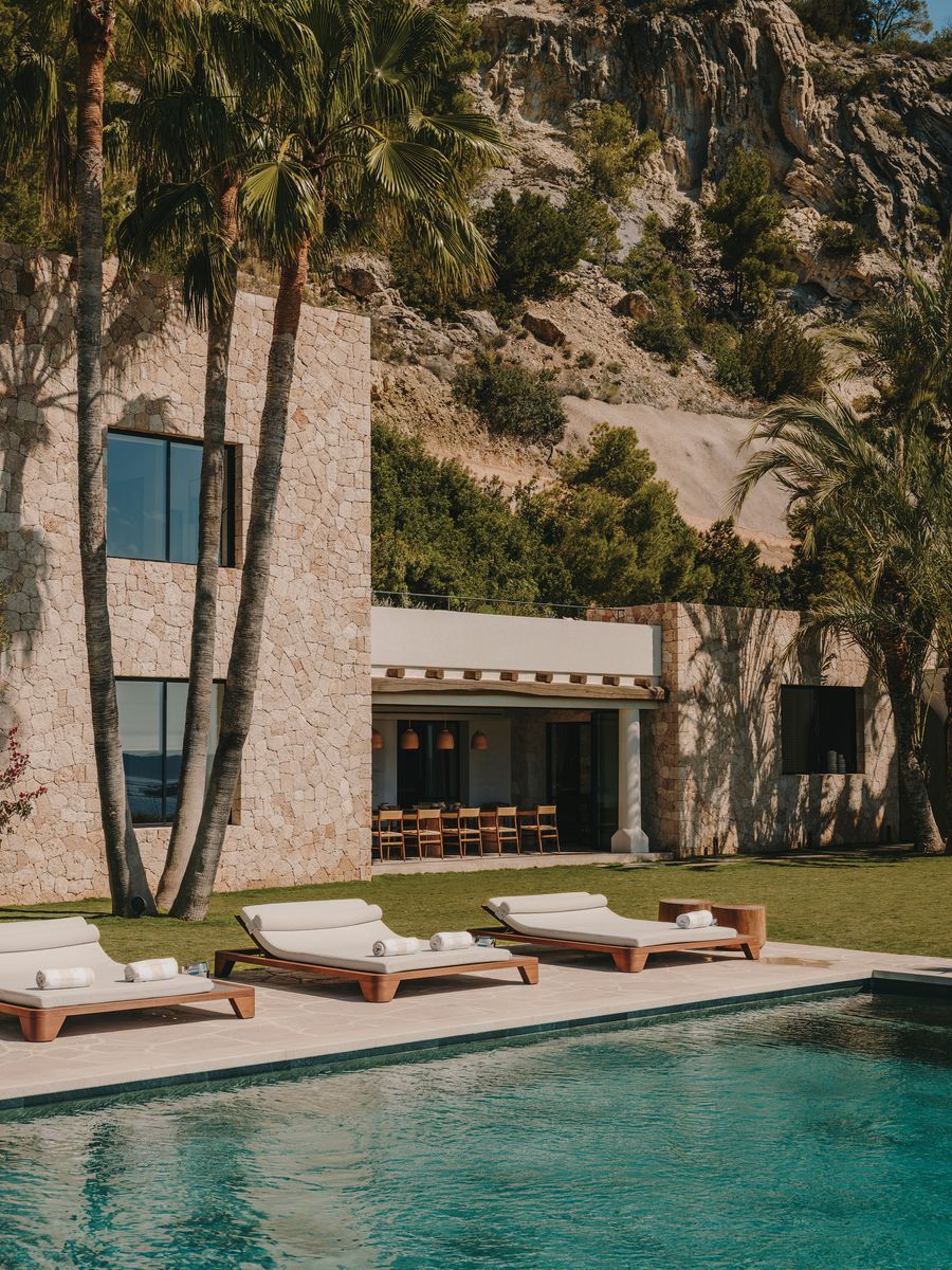 Luxurious villa with a stone facade, poolside lounge chairs, surrounded by palm trees and rocky hills in a serene landscape. Luxurious villa with a stone facade, poolside lounge chairs, surrounded by palm trees and rocky hills in a serene landscape.