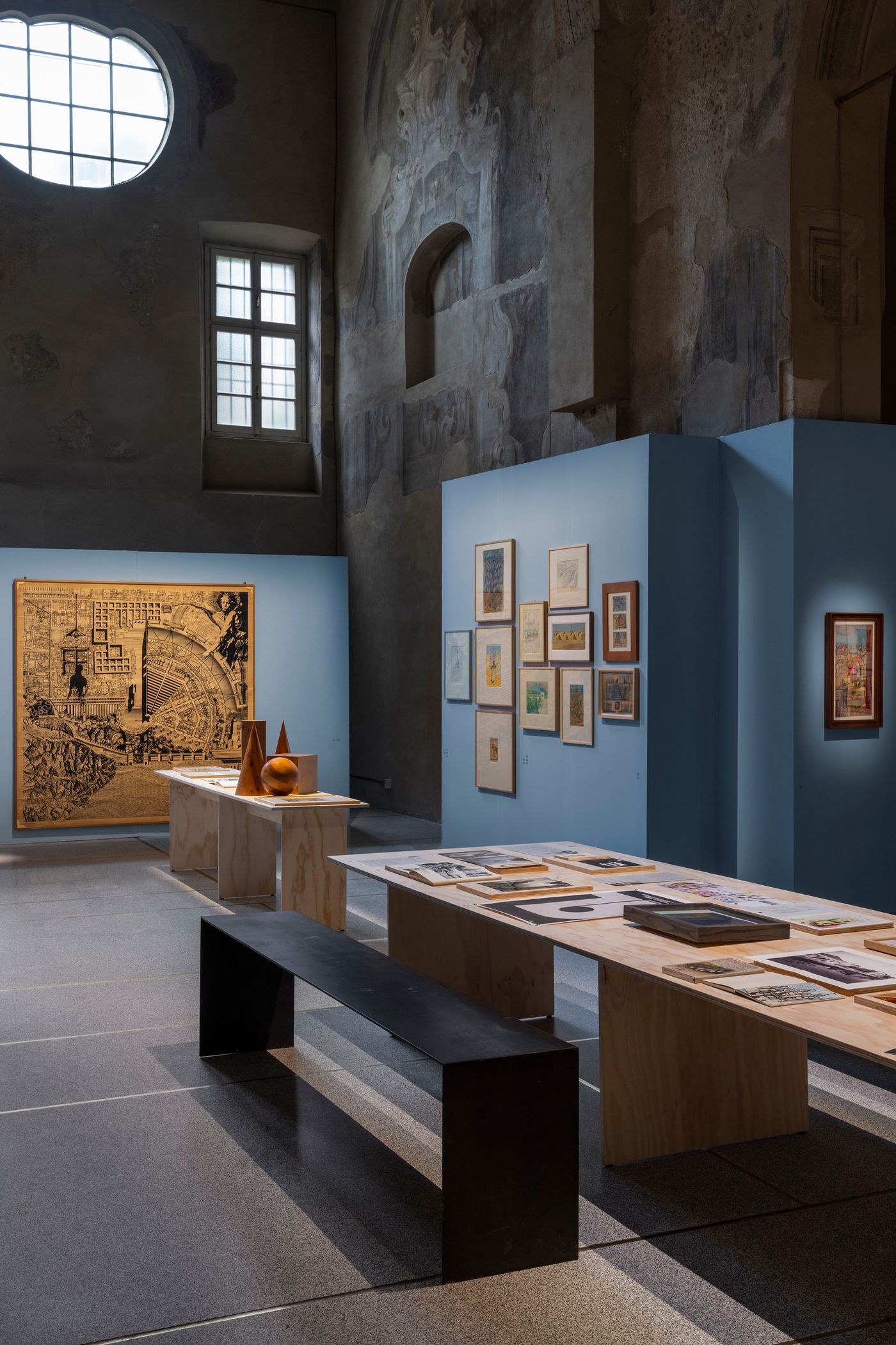 A grand hall with a high, arched ceiling and a circular window features a large, intricate gold-toned artwork with a map-like design. On wooden tables in front, framed prints and sculptural objects are displayed. The light blue exhibition walls provide a clean backdrop for a collection of smaller framed artworks, creating a focal point within the spacious, historic setting. This scene blends monumental art with curated display.
