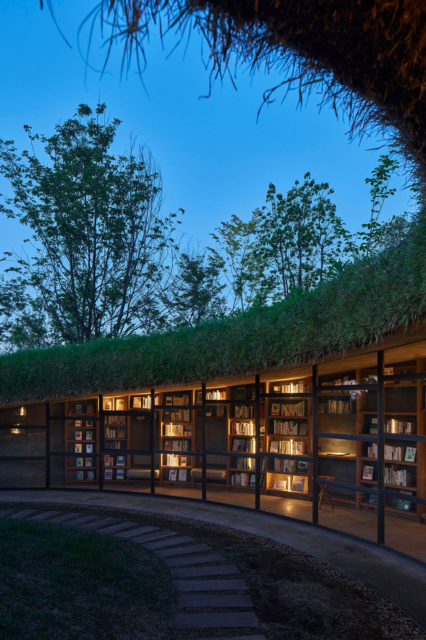 A cozy library with glass walls, illuminated bookshelves, and a grassy roof, set against a twilight sky and surrounding trees. A cozy library with glass walls, illuminated bookshelves, and a grassy roof, set against a twilight sky and surrounding trees.
