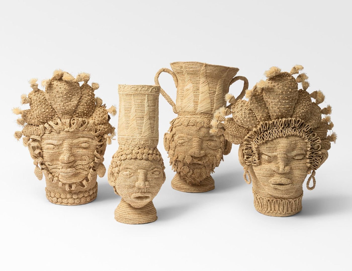 Four distinctive sculptural pieces from Natalia Brilli Studio's "Utile Collection" are presented against a clean white background. Crafted from natural fibers, possibly rattan or straw, each piece features intricate weaving that forms stylized faces and decorative headpieces reminiscent of tribal art or ancient artifacts. Their organic textures and warm, earthy tones create an authentic, artful display.