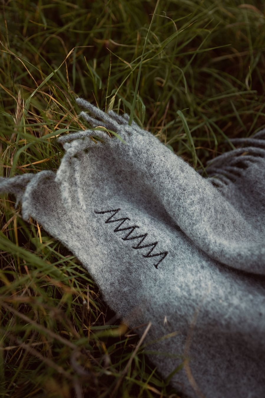 A close-up view of a grey fringed wool blanket lying on lush green grass embroidered with the MANNA hotel logo. The simple, organic stitching detail on the blanket suggests a handcrafted touch and an appreciation for natural textures, inviting a sense of comfort and connection to the outdoors. A close-up view of a grey fringed wool blanket lying on lush green grass embroidered with the MANNA hotel logo. The simple, organic stitching detail on the blanket suggests a handcrafted touch and an appreciation for natural textures, inviting a sense of comfort and connection to the outdoors.