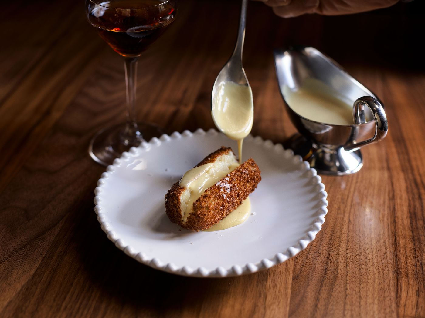 A crispy croquette is drizzled with cream sauce on a plate, with a glass in the background on a wooden table. A crispy croquette is drizzled with cream sauce on a plate, with a glass in the background on a wooden table.