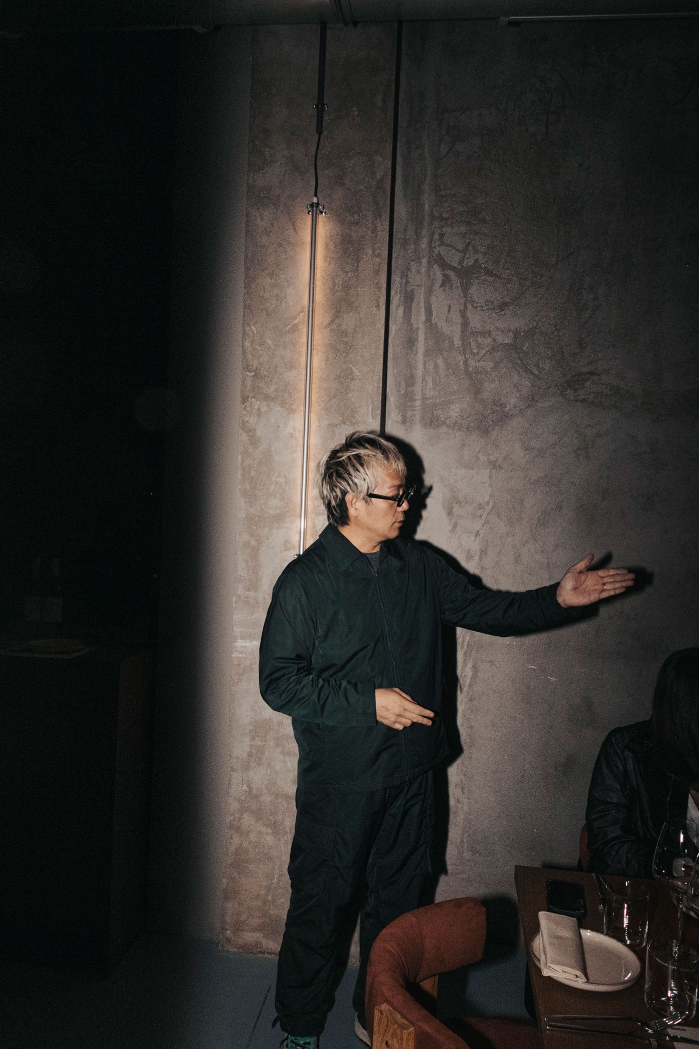 A person in dark clothing gestures while standing near a bare concrete wall, illuminated by a light fixture overhead.