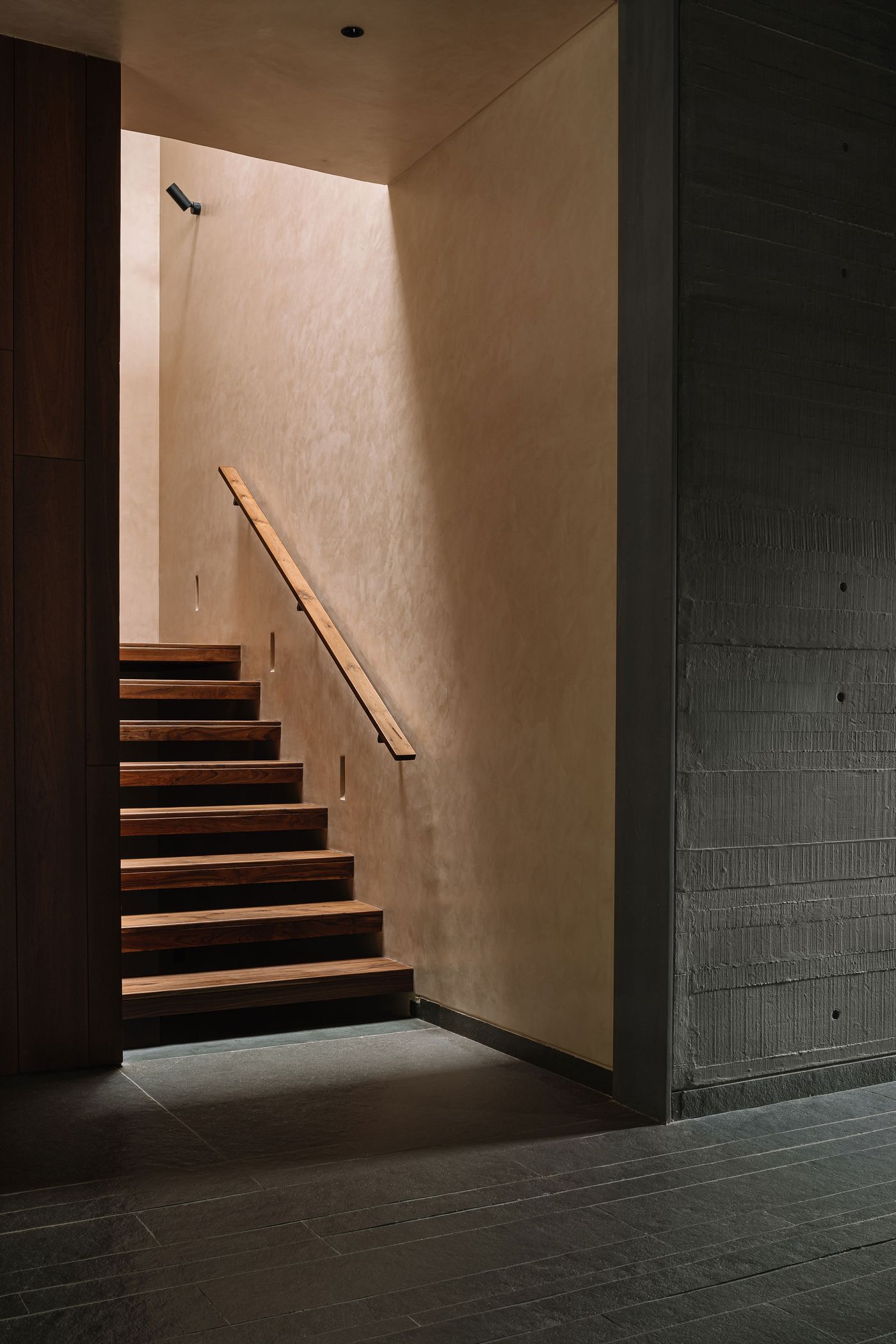 A dramatic staircase with warm wooden treads illuminated by natural light filtering down from above, creating striking shadows on the soft-toned plaster wall. A sleek wooden handrail provides a minimalist guide. The raw, board-formed concrete wall on the right adds a textural contrast, highlighting the interplay of light, shadow, and materials.
