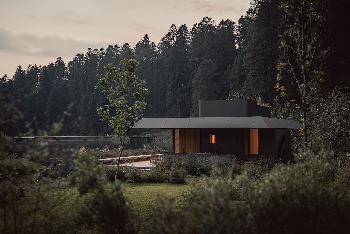 Modern dark-hued house nestled by a serene lake, surrounded by lush trees and greenery at dusk, reflecting a tranquil atmosphere.