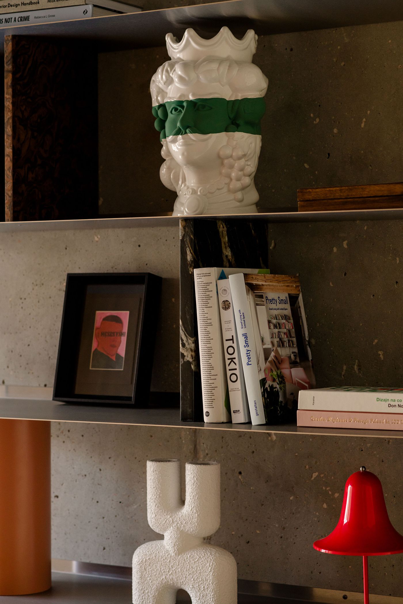 A closer look at the bespoke shelving reveals a theatrical arrangement of bold textures and curiosities—from a green-masked ceramic bust to architectural books and Memphis-style objects, all layered against a raw concrete backdrop.