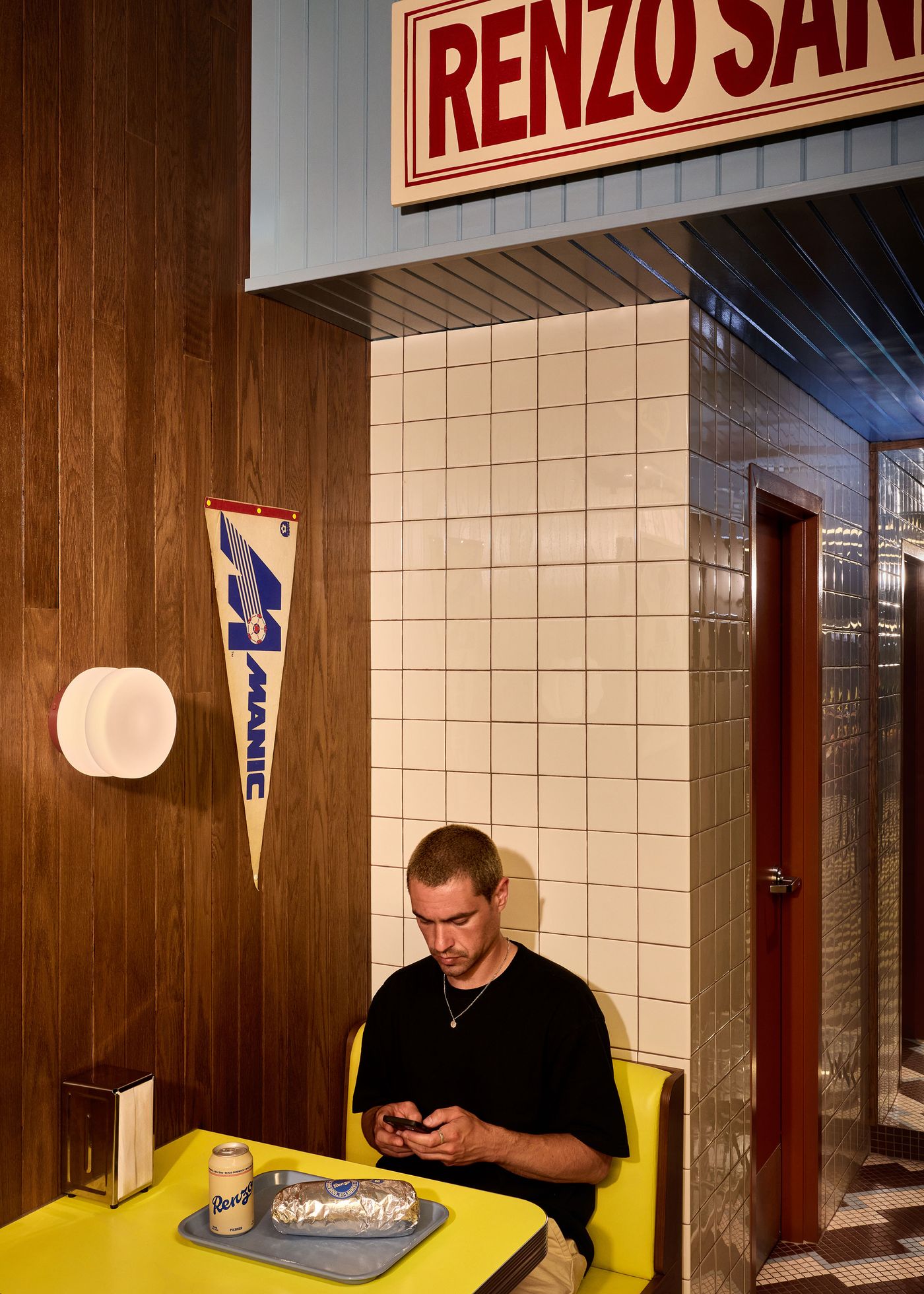 A young man sits in a yellow booth at Renzo Sandwich Shop, engrossed in his phone. A can of "Renzo" soda and a foil-wrapped sandwich rest on the table before him. The retro interior, with its wood paneling and a vintage "MANIC" pennant, creates a casual and nostalgic atmosphere.