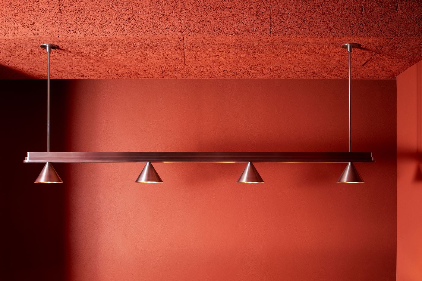 A striking linear pendant light fixture featuring four silver cone-shaped lamps, suspended against a bold, terracotta-colored wall. The minimalist design of the fixture provides a clean, modern contrast to the richly textured wall and ceiling, highlighting Naessi Studio's refined approach to lighting within Santo Palato's inviting ambiance. A striking linear pendant light fixture featuring four silver cone-shaped lamps, suspended against a bold, terracotta-colored wall. The minimalist design of the fixture provides a clean, modern contrast to the richly textured wall and ceiling, highlighting Naessi Studio's refined approach to lighting within Santo Palato's inviting ambiance.