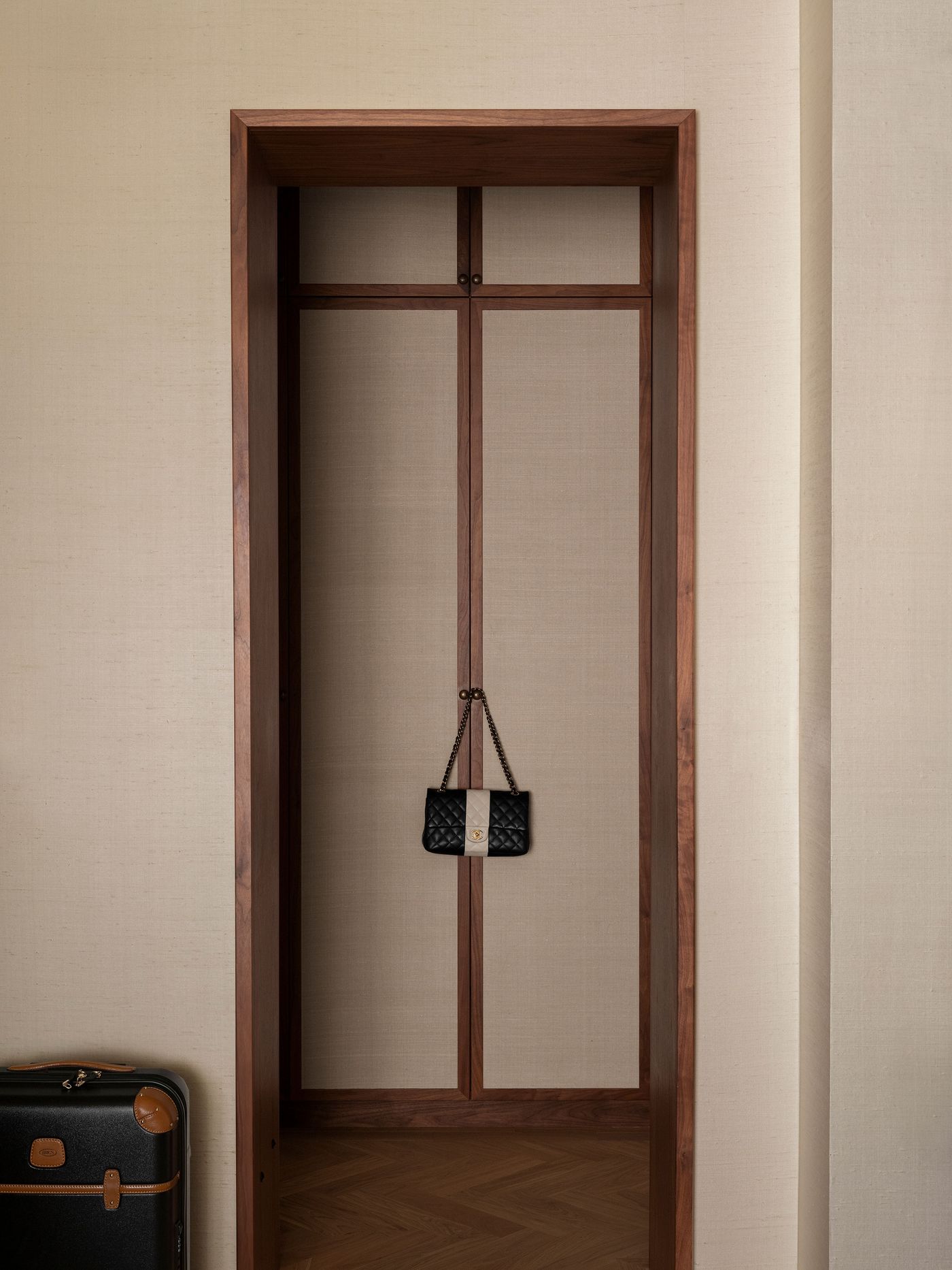 A minimalist entryway features a wooden frame with a beige backdrop, displaying a black quilted handbag and a vintage suitcase nearby. A minimalist entryway features a wooden frame with a beige backdrop, displaying a black quilted handbag and a vintage suitcase nearby.