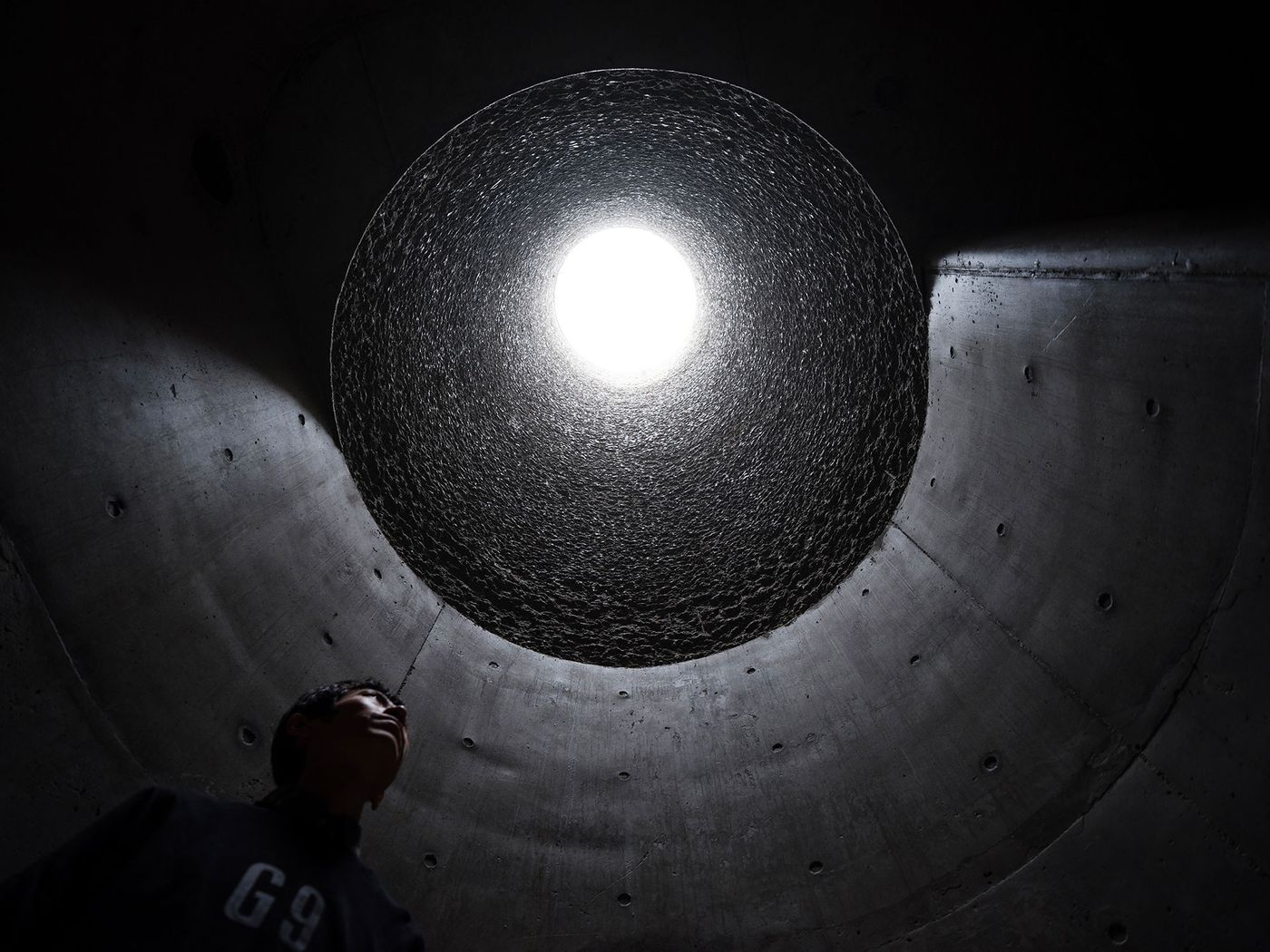 A person stands in a dark, cylindrical space, gazing up at a bright circular light source above. A person stands in a dark, cylindrical space, gazing up at a bright circular light source above.