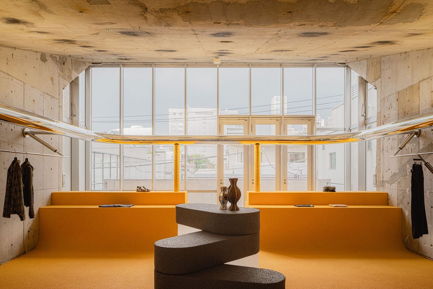 The upper floor opens towards a broad glazed façade, with canary-yellow microcement rising into low perimeter benches and display platforms. At centre, a stacked sculptural counter anchors the space, while chrome rails sweep along the raw concrete walls, creating a precise metallic line through I IN’s deliberately unfinished setting.