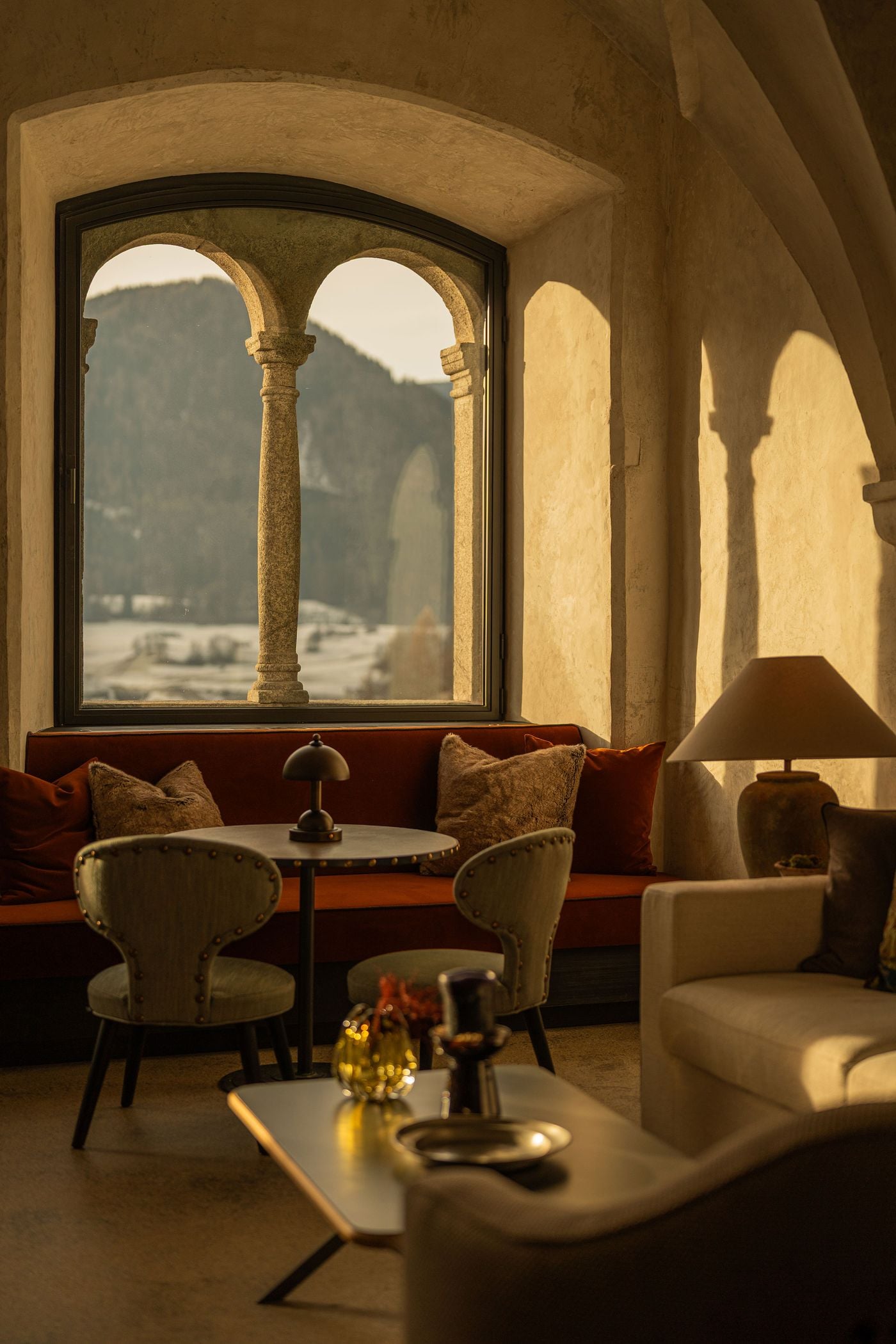 Framed by modern steel glazing, a medieval twin-arched window with a slender granite column looks out over snowy fields and forested slopes from Castel Badia's vaulted lounge. A domed lamp, studded chairs and a velvet banquette fill the deep window bay, while the arches cast soft shadows on the wall. Framed by modern steel glazing, a medieval twin-arched window with a slender granite column looks out over snowy fields and forested slopes from Castel Badia's vaulted lounge. A domed lamp, studded chairs and a velvet banquette fill the deep window bay, while the arches cast soft shadows on the wall.