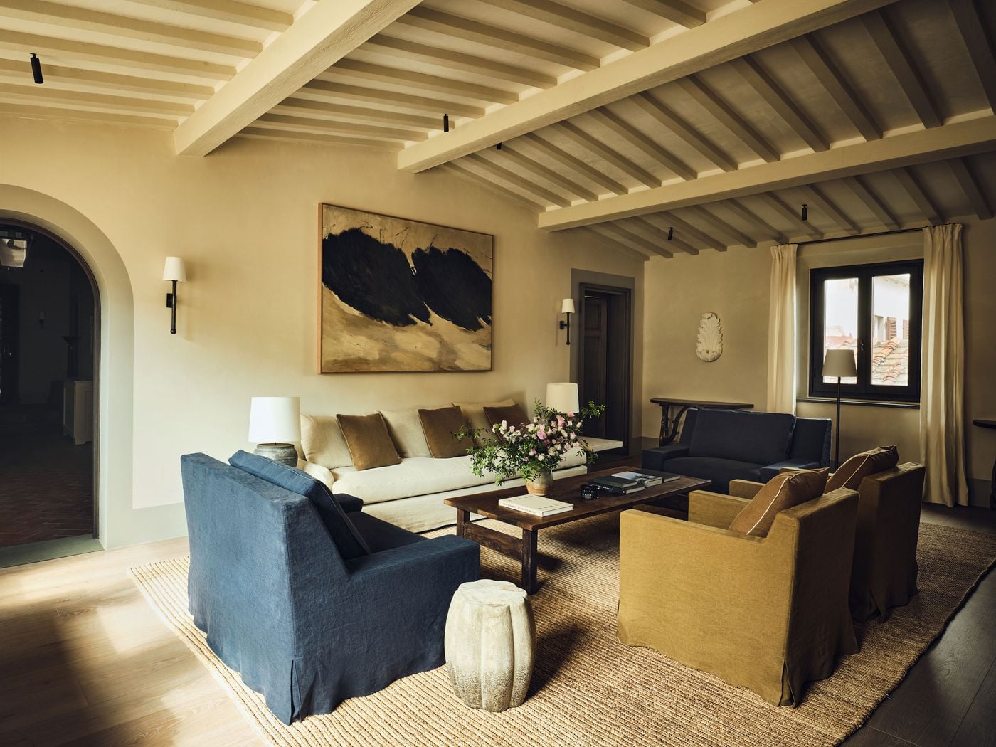 Pale limewashed timber beams span a top-floor living room at La Réserve Firenze, where a large abstract canvas of black brushstrokes on ochre presides over a linen sofa. Slipcovered armchairs in indigo and mustard, a rustic coffee table, a jute rug and a window onto terracotta rooftops give the room a relaxed, collected warmth. Pale limewashed timber beams span a top-floor living room at La Réserve Firenze, where a large abstract canvas of black brushstrokes on ochre presides over a linen sofa. Slipcovered armchairs in indigo and mustard, a rustic coffee table, a jute rug and a window onto terracotta rooftops give the room a relaxed, collected warmth.