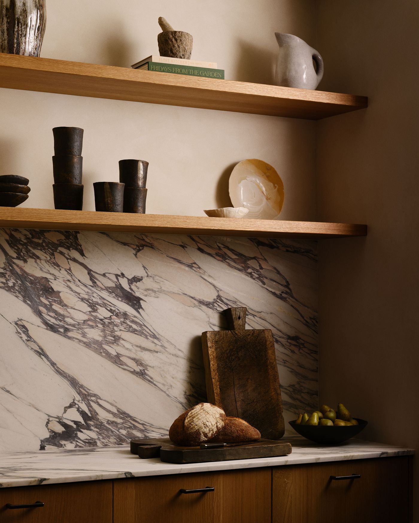A rustic kitchen scene featuring marble countertops, wooden shelves with pottery, and a cutting board with fresh bread and pears.