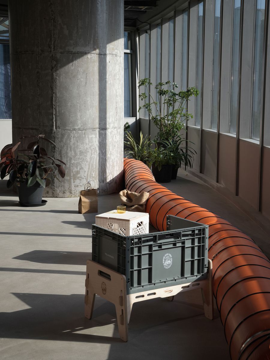 This intriguing corner of 889 GLO Art Space features a raw concrete pillar contrasting with an exposed orange ventilation tube, a bold industrial element. Potted plants introduce biophilic accents, while an innovative, modular seating design using crates on a wooden base highlights the space's experimental and resourceful design philosophy. This intriguing corner of 889 GLO Art Space features a raw concrete pillar contrasting with an exposed orange ventilation tube, a bold industrial element. Potted plants introduce biophilic accents, while an innovative, modular seating design using crates on a wooden base highlights the space's experimental and resourceful design philosophy.