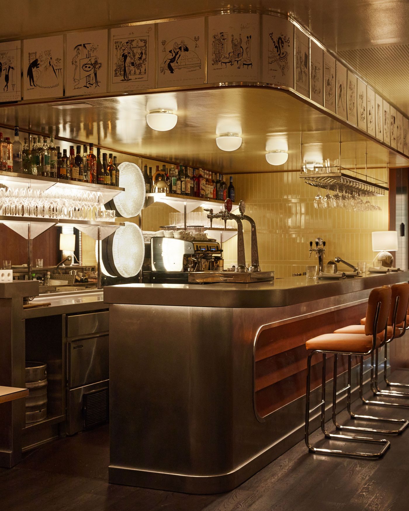 A full view of a vibrant bar area with a strong Art Deco influence. The curved, stainless steel counter is a striking focal point, complemented by low-slung, tan leather bar stools. The space is crowned by a line of whimsical, circular ceiling lights and a frieze of framed drawings, creating an elegant and playful environment. A full view of a vibrant bar area with a strong Art Deco influence. The curved, stainless steel counter is a striking focal point, complemented by low-slung, tan leather bar stools. The space is crowned by a line of whimsical, circular ceiling lights and a frieze of framed drawings, creating an elegant and playful environment.