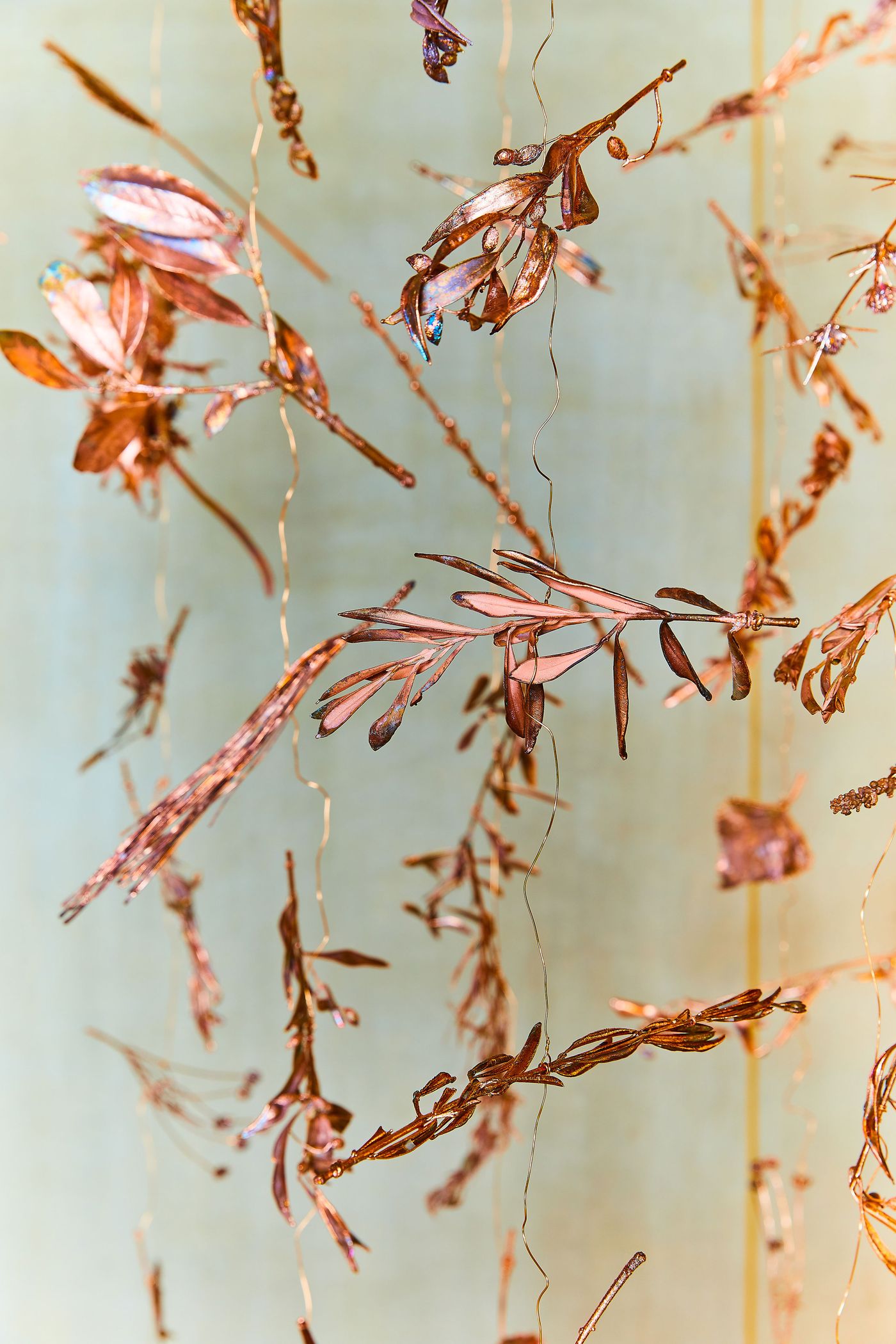 An intimate view of Gabriel Leger's suspended installation, showing a collection of small, biomorphic forms crafted from gilded metal and thin wire. The light illuminates the pieces, creating a soft, warm glow that highlights the intricate details of the leaves and stems. The abstract, airy composition contrasts with the solid, traditional space.