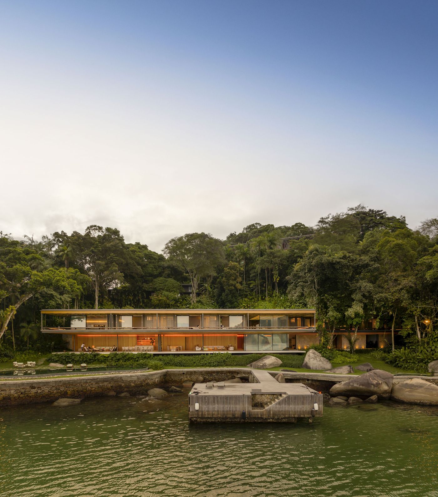 The modernist coastal dwelling glows with interior light at dusk, its horizontal layers of warm wood and glass nestled against a dense backdrop of tropical forest. A private timber dock extends over the calm, green-tinged waters, enhancing the serene and inviting atmosphere of this integrated architectural gem. The modernist coastal dwelling glows with interior light at dusk, its horizontal layers of warm wood and glass nestled against a dense backdrop of tropical forest. A private timber dock extends over the calm, green-tinged waters, enhancing the serene and inviting atmosphere of this integrated architectural gem.