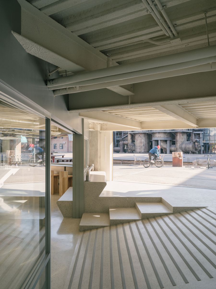 An intriguing transition space reveals a multi-level seating area composed of minimalist, light grey concrete steps, leading down to a ribbed floor texture. Through expansive glass, an industrial outdoor setting is visible, with a person on a bicycle, showcasing the seamless blend of interior design with the external environment, and demonstrating biophilic principles. An intriguing transition space reveals a multi-level seating area composed of minimalist, light grey concrete steps, leading down to a ribbed floor texture. Through expansive glass, an industrial outdoor setting is visible, with a person on a bicycle, showcasing the seamless blend of interior design with the external environment, and demonstrating biophilic principles.