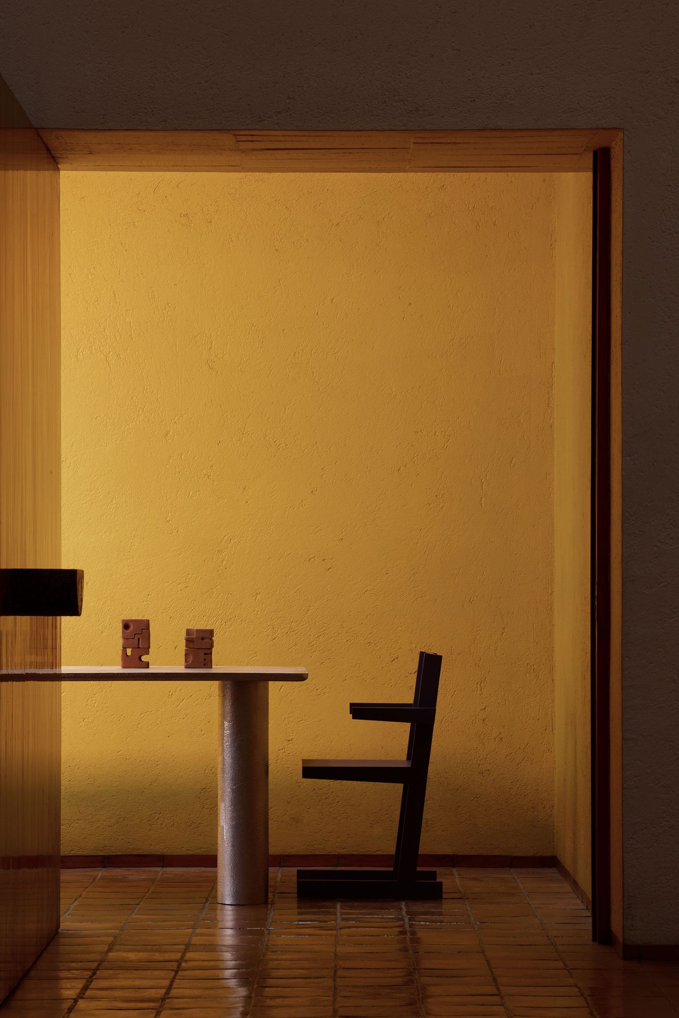 A minimalist dining space features a cylindrical table, a dark wooden chair, and two small sculptures, illuminated by a warm yellow wall.