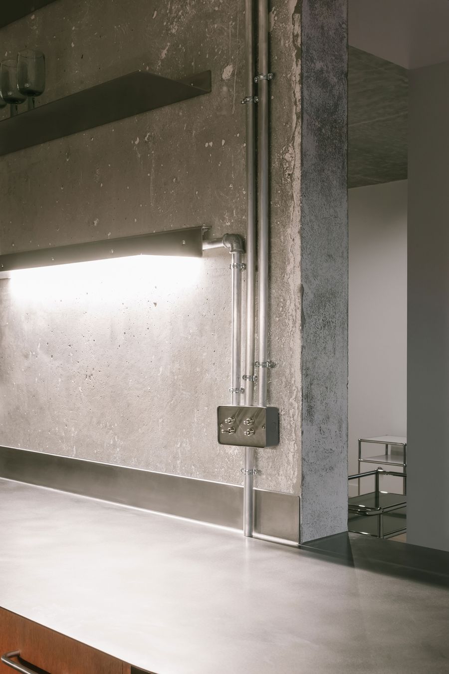 A modern kitchen corner featuring a concrete wall, metallic pipes, overhead lighting, and a sleek stainless steel countertop. A modern kitchen corner featuring a concrete wall, metallic pipes, overhead lighting, and a sleek stainless steel countertop.