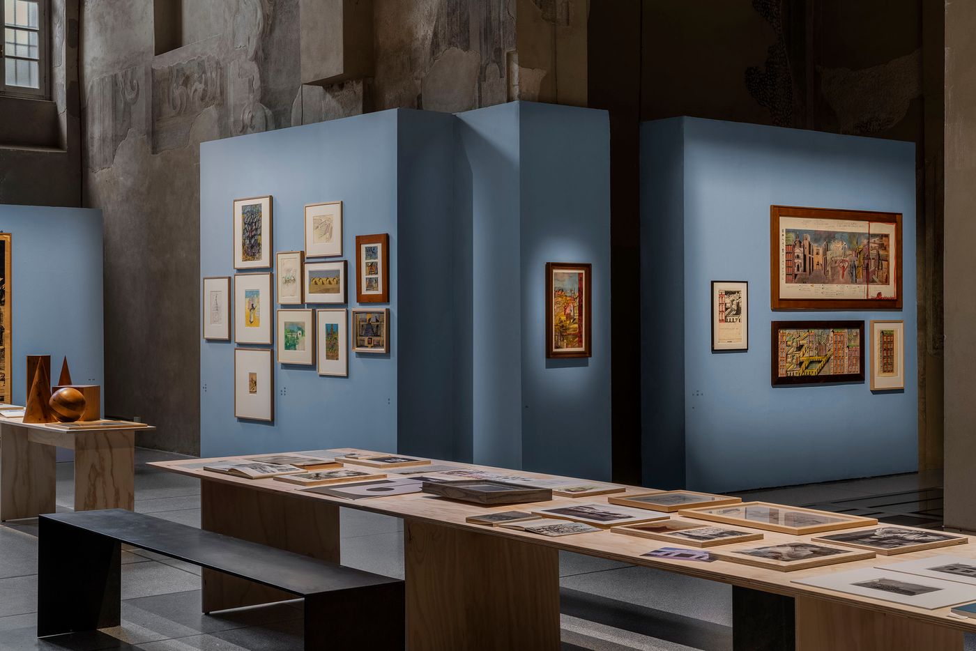Within a hall with weathered, frescoed walls, minimalist light blue exhibition walls create distinct display zones. A long wooden table is laid out with numerous framed prints and books, inviting close inspection. Other displays feature framed artworks of varying sizes, some with intricate urban or landscape scenes, arranged on these partitions. The overall presentation is clean and contemporary, highlighting the exhibited works against the historical backdrop.