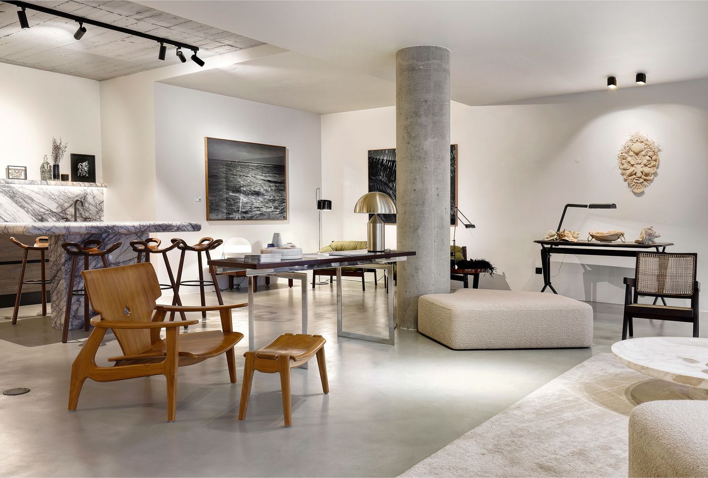 A sophisticated showroom interior from Estadeobruto, part of Lisbon Design Week, blends brutalist and mid-century modern elements. A raw concrete column anchors the open space, complemented by a dramatic veined marble bar. Warm wooden seating, a minimalist metal-framed table, and ambient track lighting highlight the curated collection of art and furniture, creating an atmosphere of refined industrial elegance. A sophisticated showroom interior from Estadeobruto, part of Lisbon Design Week, blends brutalist and mid-century modern elements. A raw concrete column anchors the open space, complemented by a dramatic veined marble bar. Warm wooden seating, a minimalist metal-framed table, and ambient track lighting highlight the curated collection of art and furniture, creating an atmosphere of refined industrial elegance.