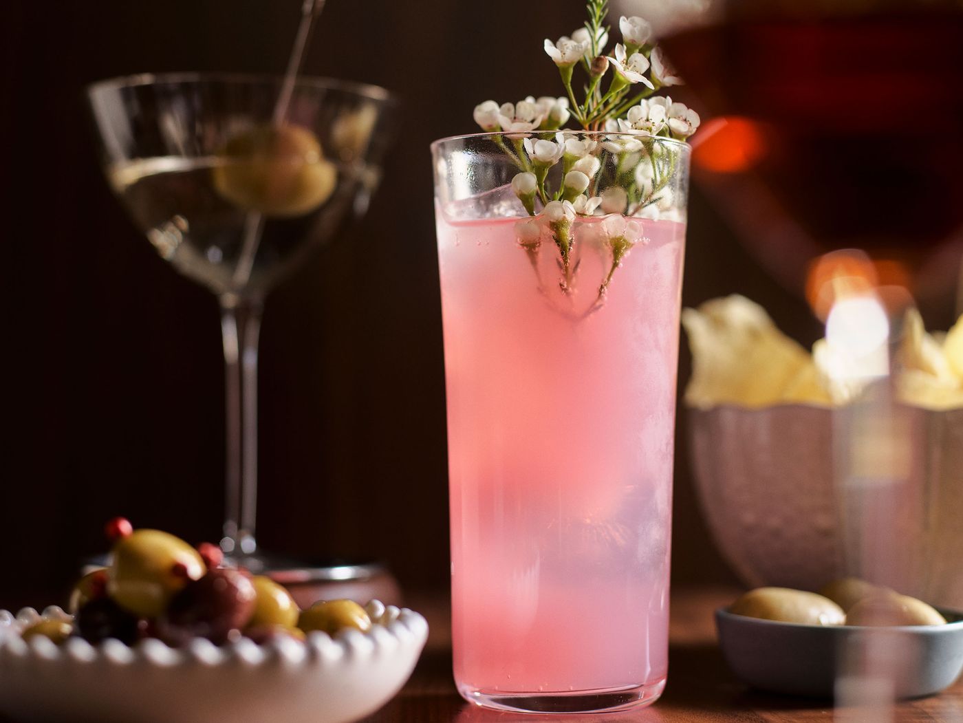 A refreshing pink cocktail garnished with delicate white flowers, alongside a martini and a bowl of olives and potato chips. A refreshing pink cocktail garnished with delicate white flowers, alongside a martini and a bowl of olives and potato chips.