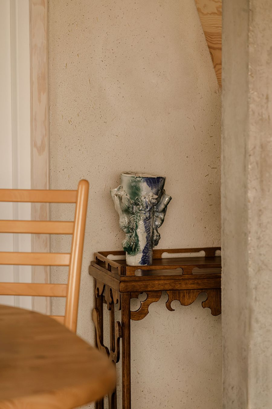 A colorful ceramic vase rests on an intricately designed wooden side table, beside a wooden chair and textured wall. A colorful ceramic vase rests on an intricately designed wooden side table, beside a wooden chair and textured wall.