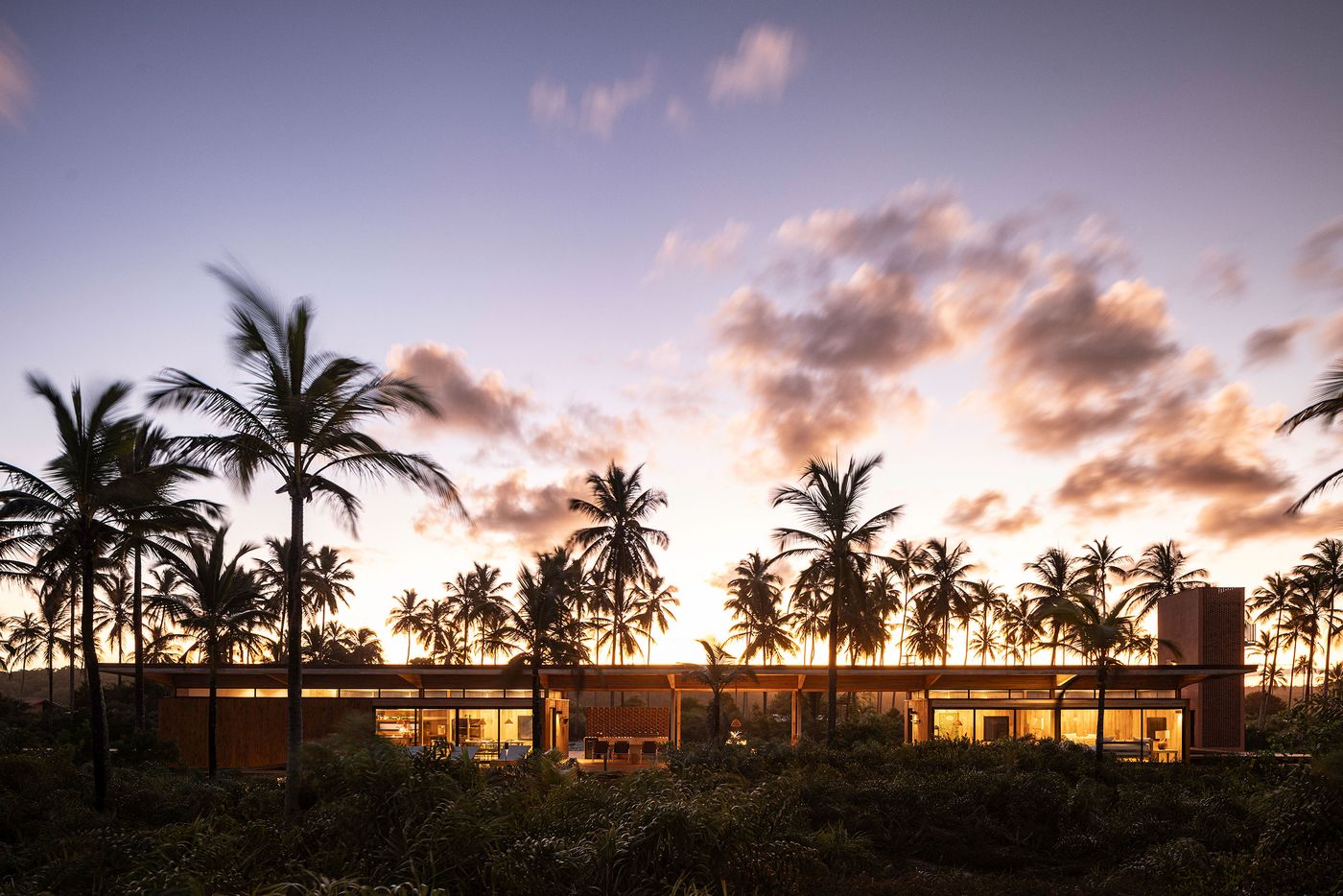 A wide shot of an softly illuminated, low-slung building, featuring extensive glass and warm timber, nestled among a dense forest of palm trees at sunset. The architectural forms blend seamlessly with the natural landscape, creating a luminous, serene tropical escape under a dramatic sky. A wide shot of an softly illuminated, low-slung building, featuring extensive glass and warm timber, nestled among a dense forest of palm trees at sunset. The architectural forms blend seamlessly with the natural landscape, creating a luminous, serene tropical escape under a dramatic sky.