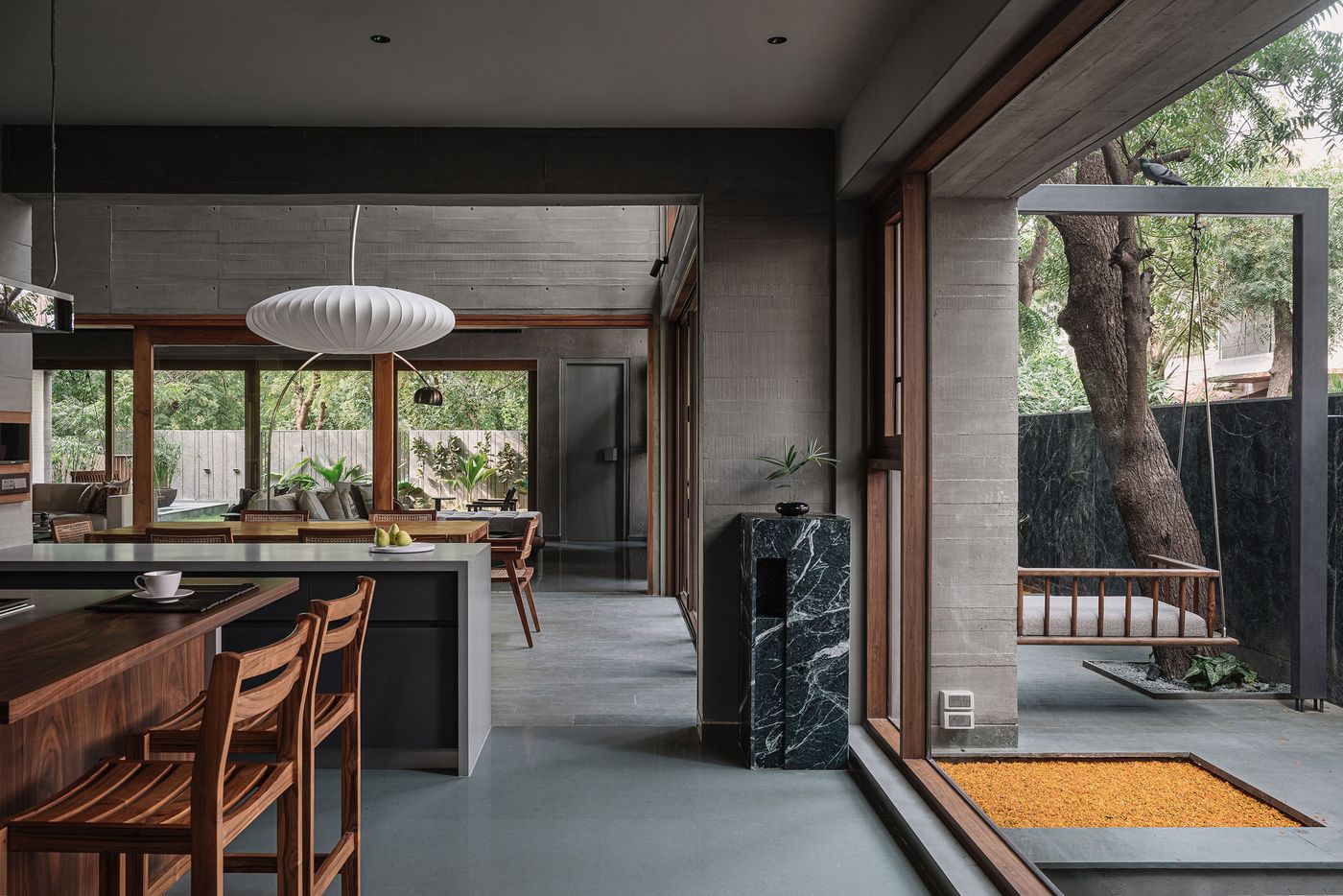 A modern open-plan living area that flows seamlessly between kitchen, dining, and outdoor spaces. Raw concrete walls and a muted floor create a minimalist backdrop. A striking sculptural light fixture hangs above a dining area. Expansive glass doors open to an outdoor patio with a wooden swing and a black marble plinth, embracing indoor-outdoor living.