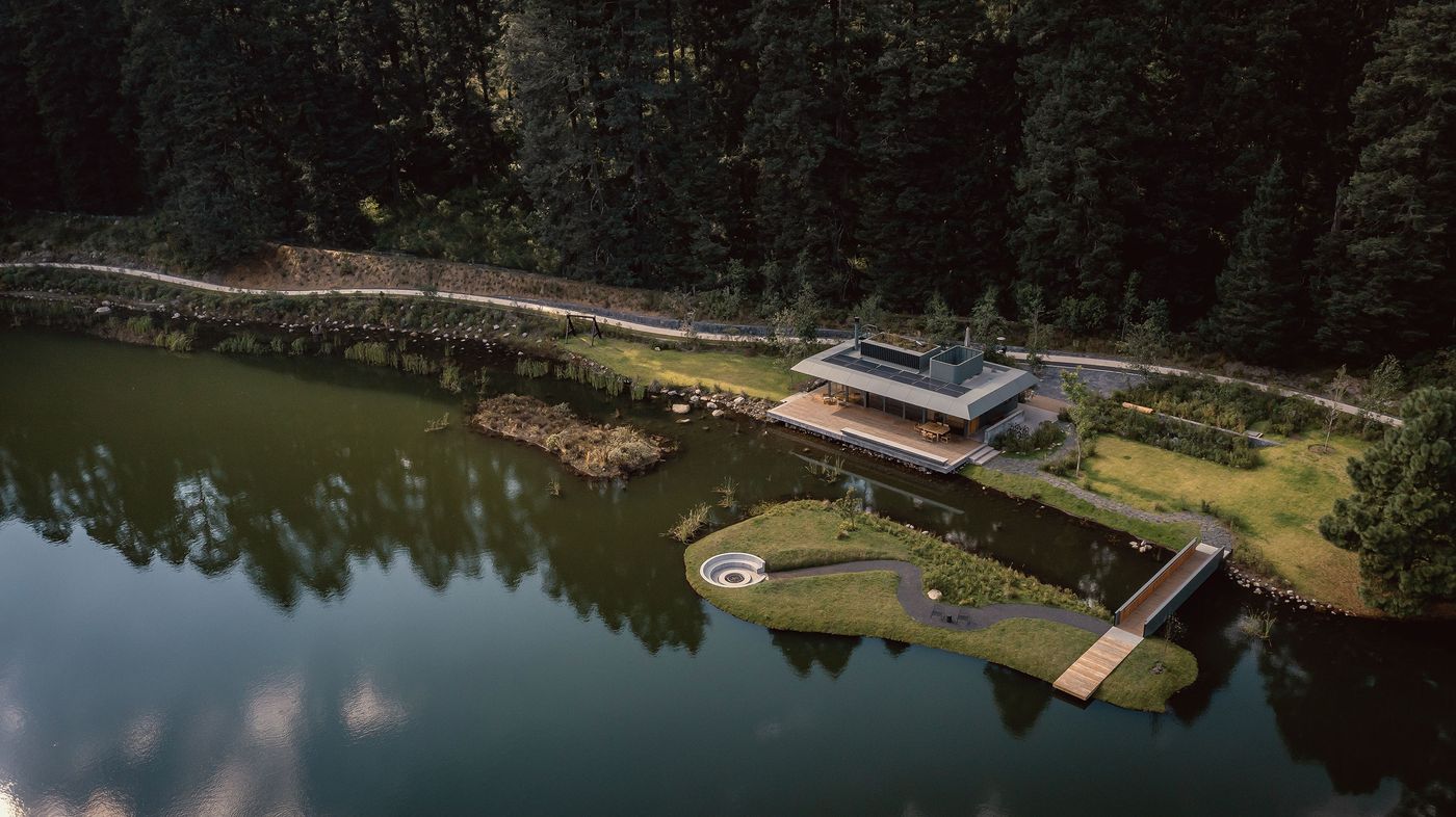 Aerial view of a modern lakeside house surrounded by trees, featuring a deck, pathway, and small island with a circular design.