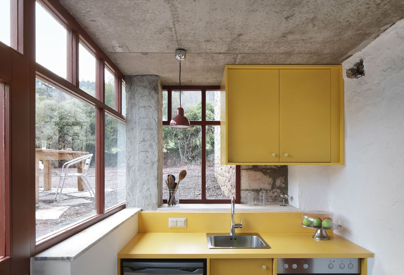 Vibrant yellow kitchen cabinetry creates a bold focal point within the transparent ground-floor pavilion, where raw concrete ceiling meets coral-red steel window frames. The compact design maximizes functionality while maintaining visual connection to the Mediterranean landscape through floor-to-ceiling glazing and thoughtful material contrasts.