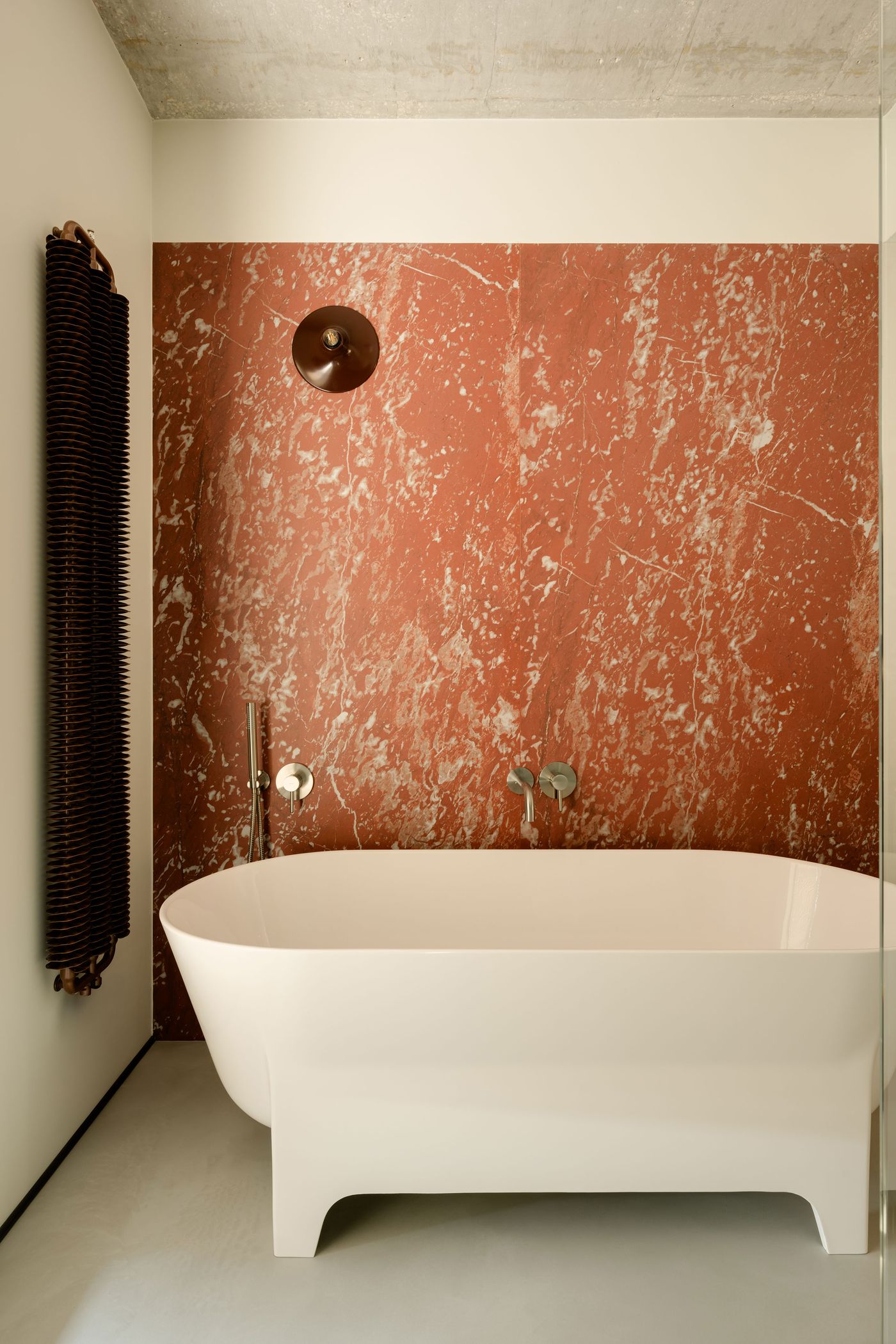 This serene bathroom showcases a sculptural white bathtub set against a striking red marble wall. The minimalist fittings and concrete ceiling balance the stone’s drama, creating a quietly luxurious bathing retreat.