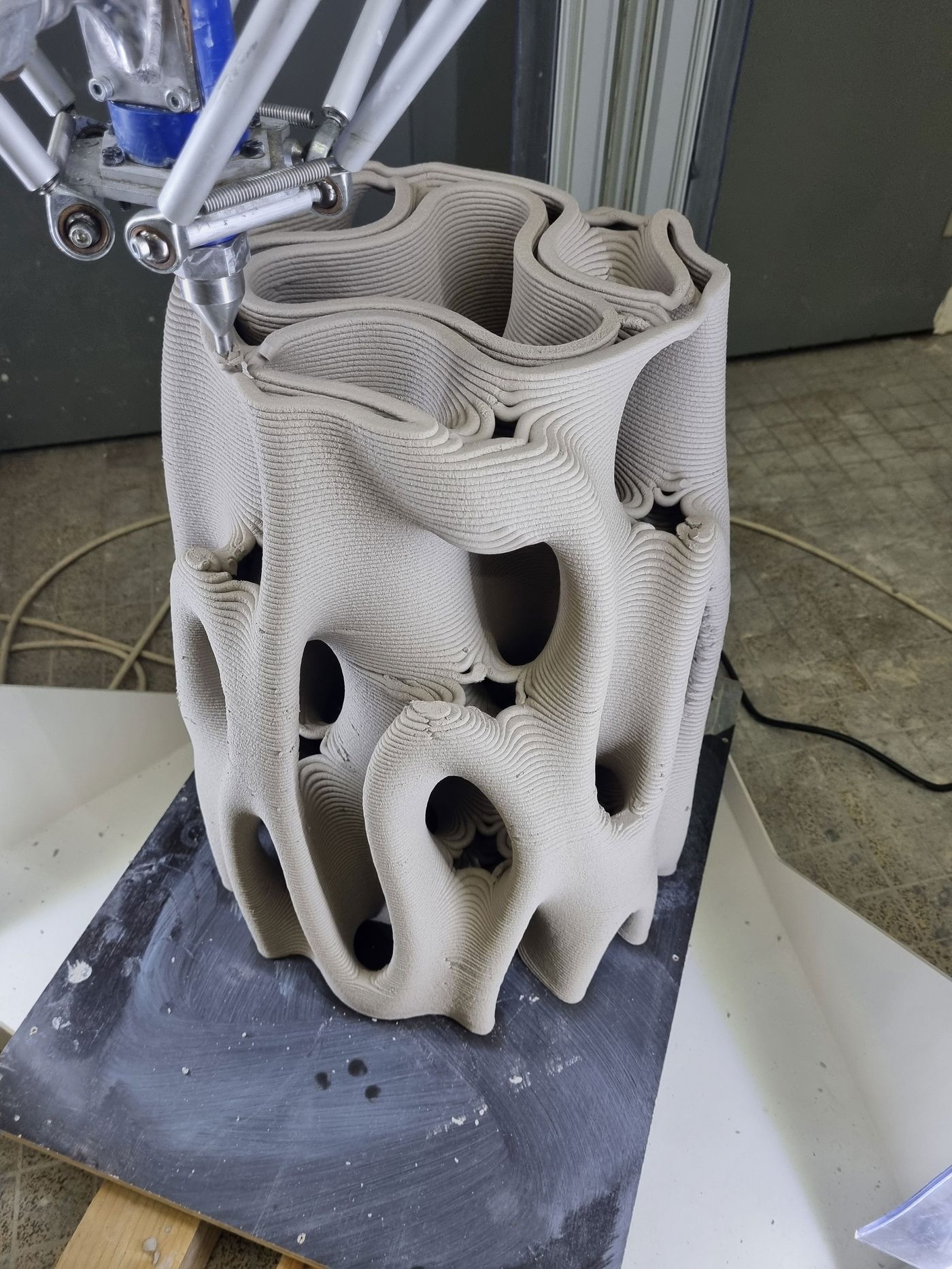 A 3D printer nozzle creating a textured, organic-shaped ceramic vase with hollow openings, resting on a base in a workshop setting.