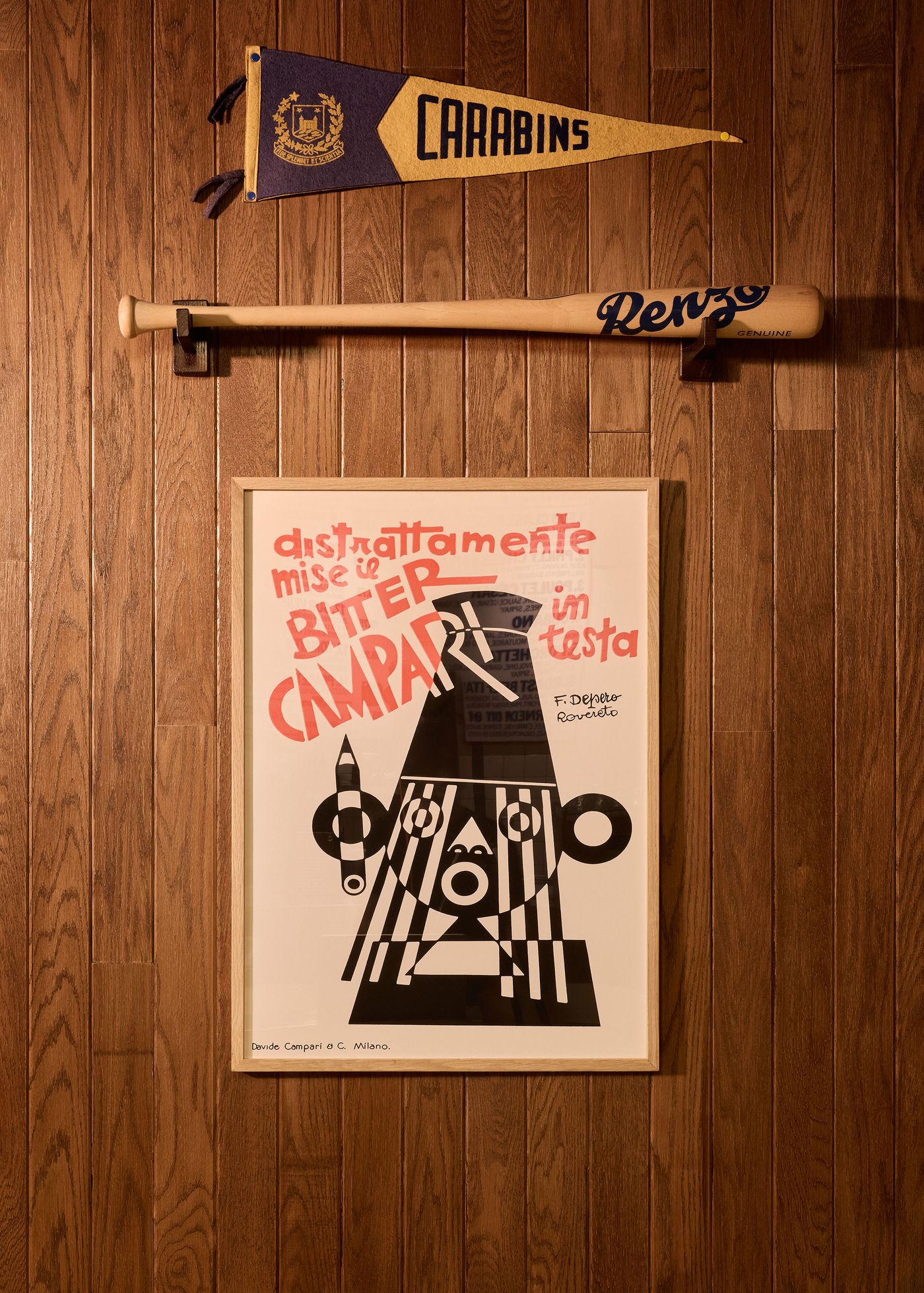 A collection of vintage-inspired decor adorns a wood-paneled wall in Renzo Sandwich. A pennant reading "CARABINS" hangs above a wooden baseball bat featuring the "Renzo" logo. Below, a framed graphic print with bold typography and abstract shapes adds an artistic, retro flair to the space.