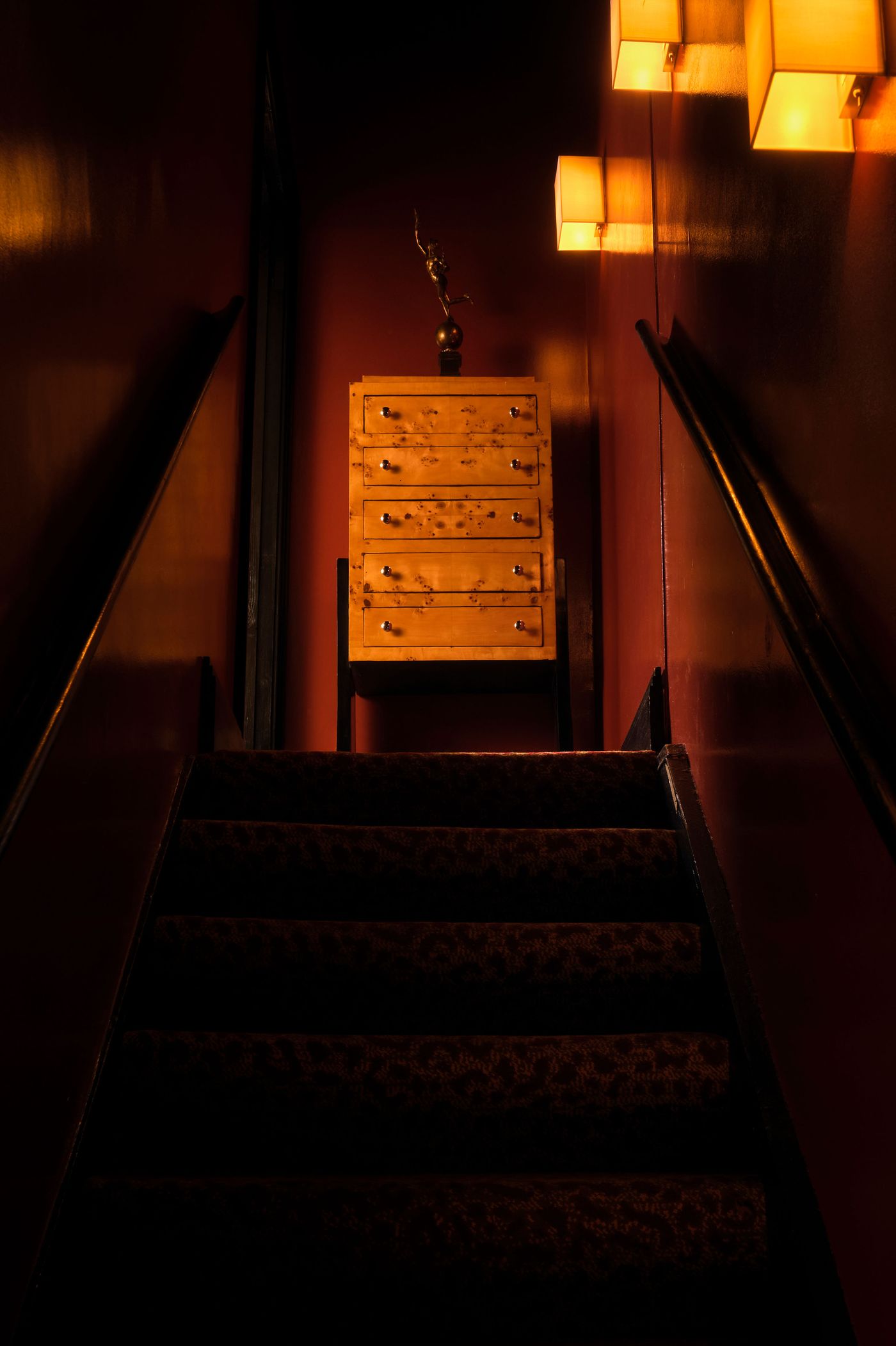 A dramatic view up a dark staircase at The Roses of Elagabalus, with deep red walls and a patterned carpet leading to a landing. At the top, a beautifully ornate wooden chest of drawers with numerous small handles stands beneath two glowing, rectangular pendant lights, contributing to the mysterious and vintage-inspired ambiance.