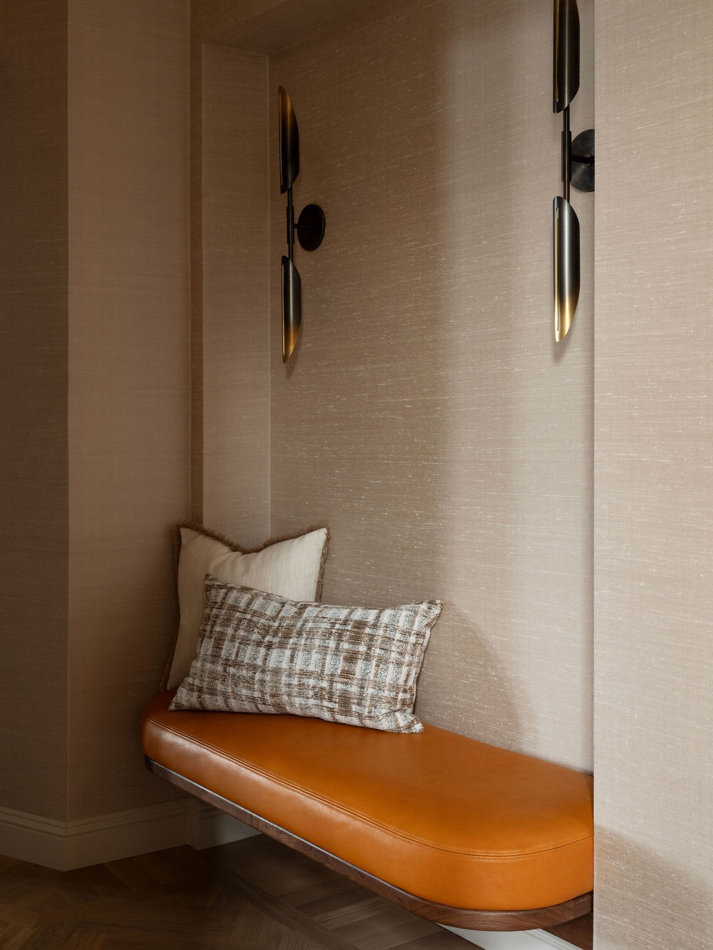 Cozy nook featuring a curved orange leather bench with decorative pillows, illuminated by stylish wall sconces against textured beige walls. Cozy nook featuring a curved orange leather bench with decorative pillows, illuminated by stylish wall sconces against textured beige walls.