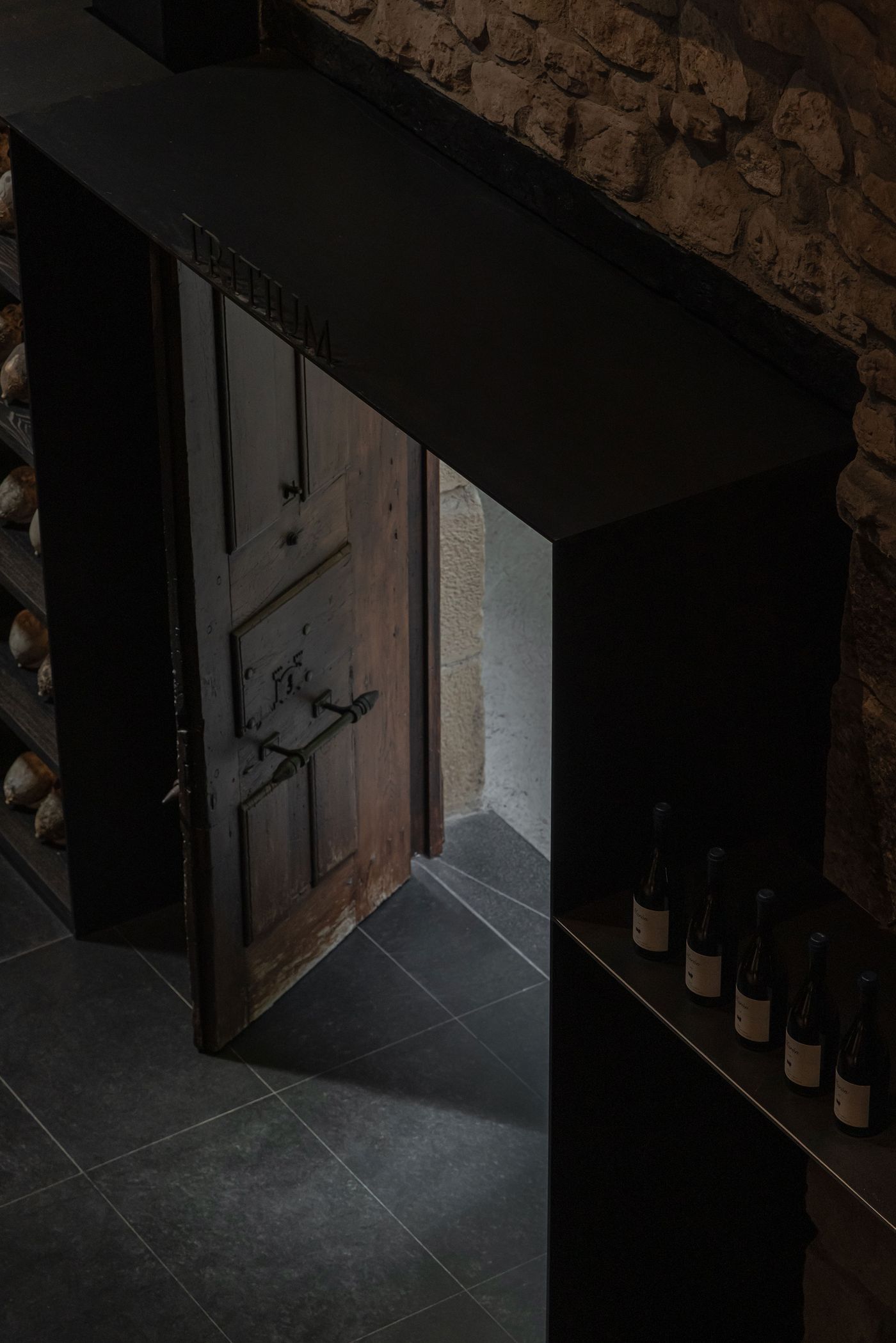 A rustic wooden door opens to a dimly lit space, framed by dark stone walls and a sleek shelf displaying wine bottles.